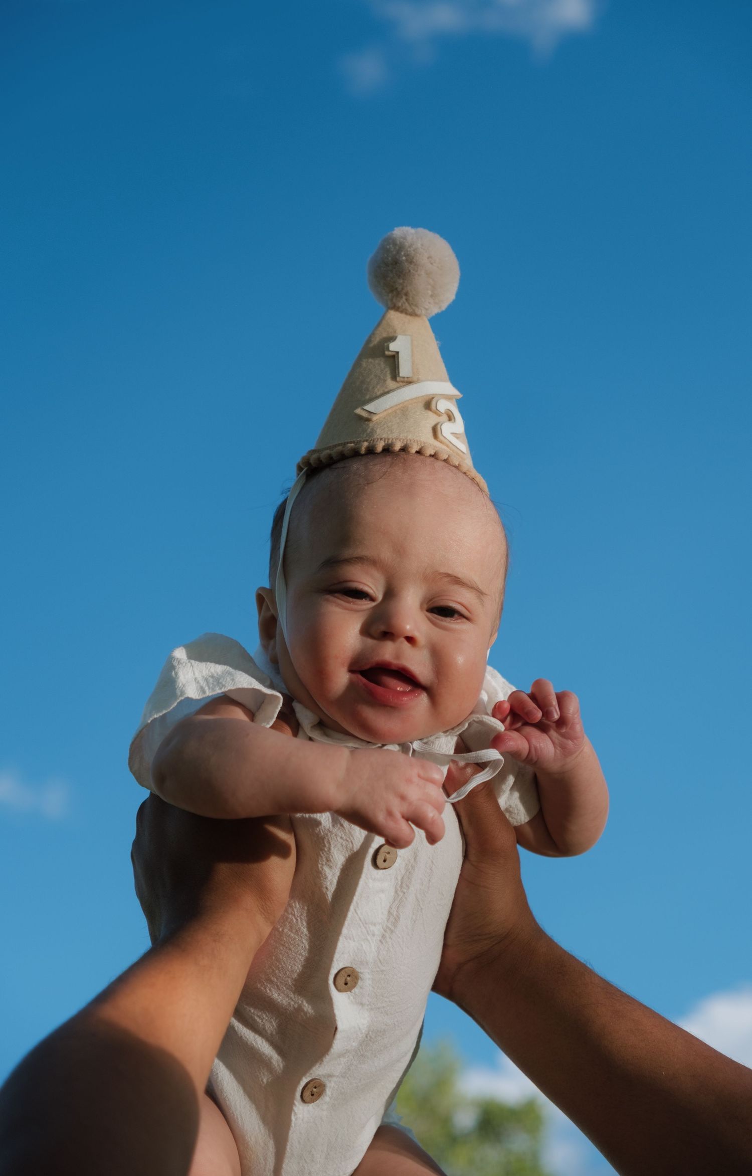 A joyful baby wearing a first birthday party hat is lifted up against a bright blue sky.