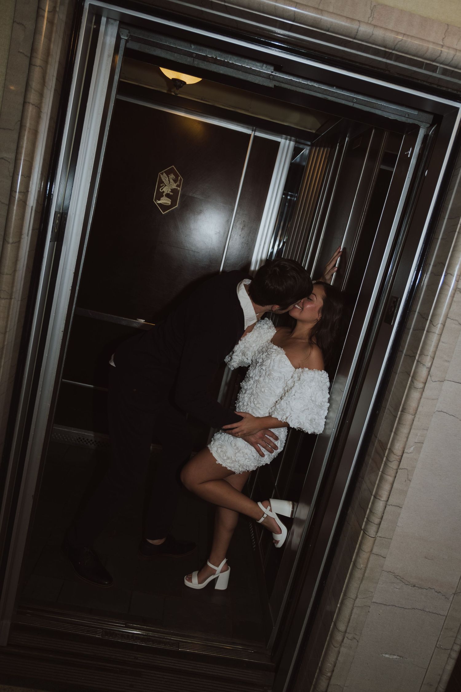 Couple sharing romantic kiss in ornate elevator, bride wearing short white lace dress and heels.