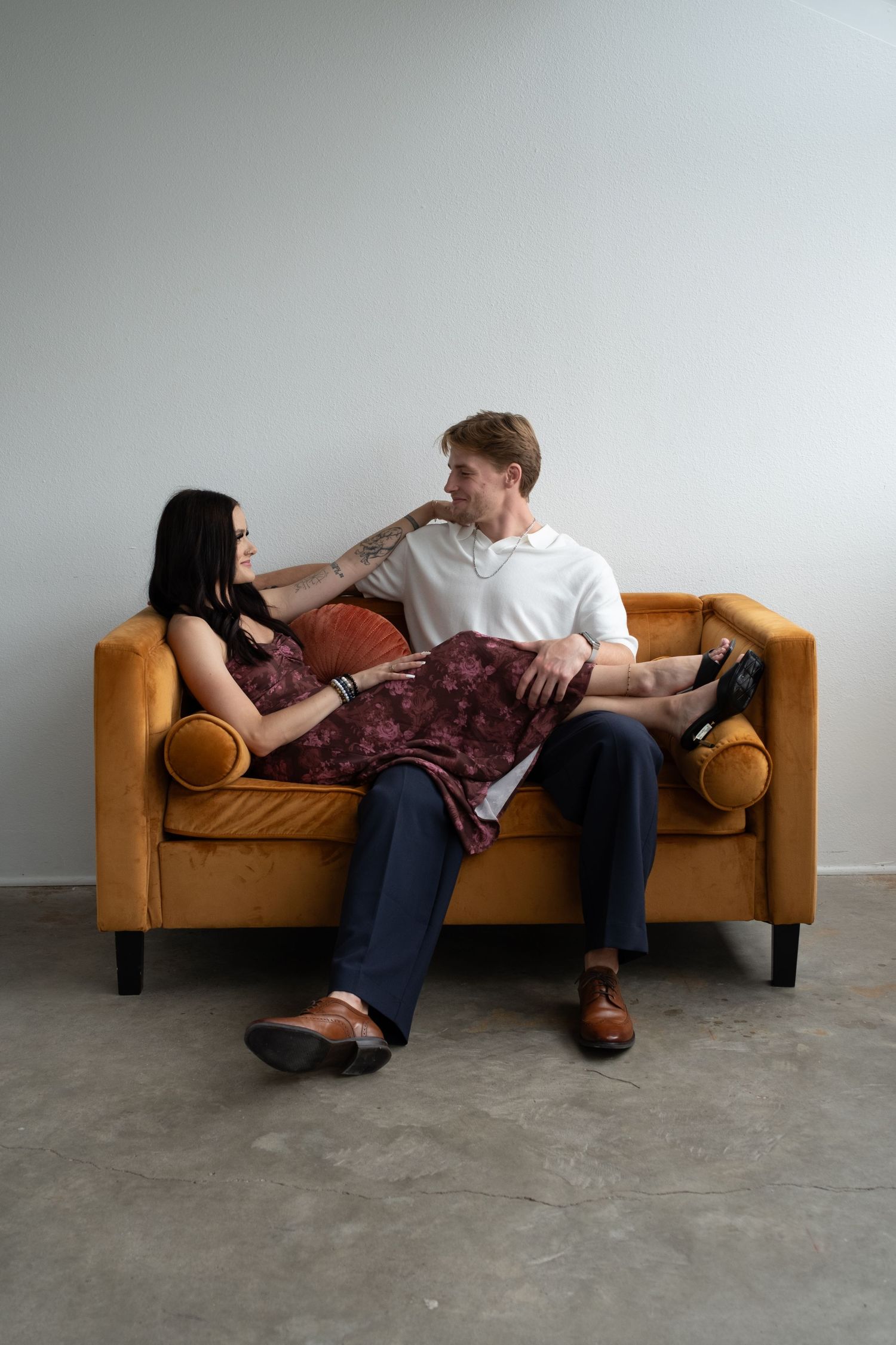 A couple relaxes on a golden velvet sofa, gazing at each other in a minimalist room with concrete floors.