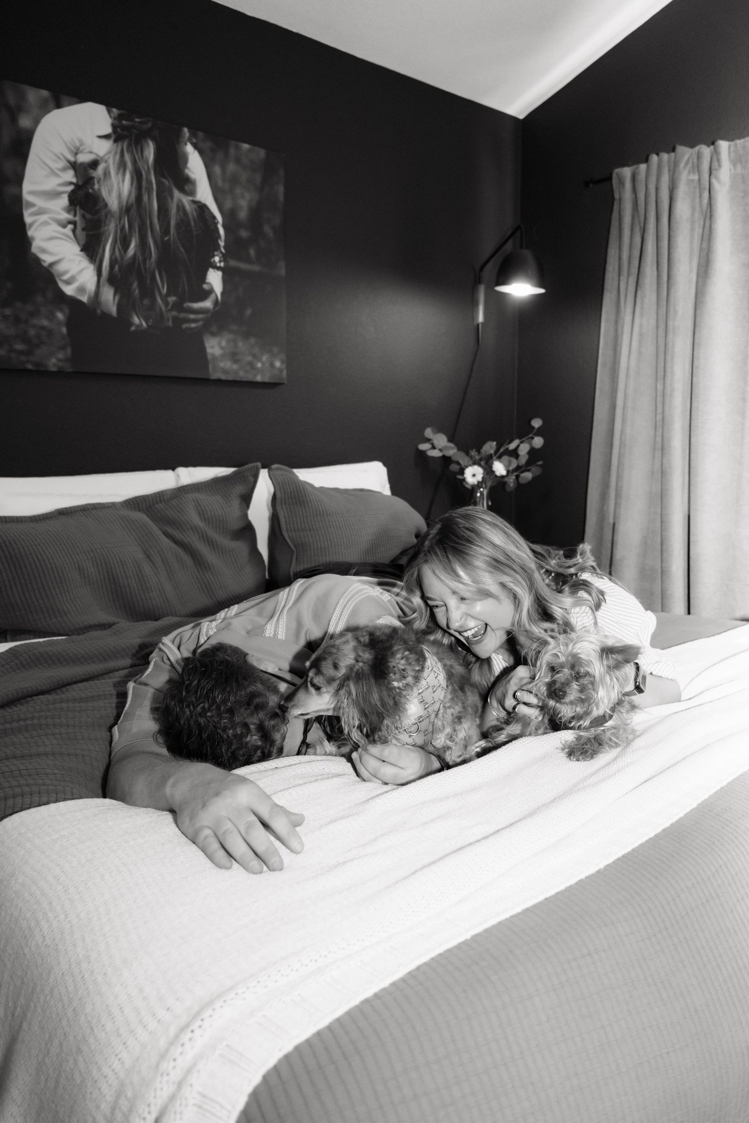 Black and white photo of a smiling woman and a man cuddling two small dogs on a cozy bed in a dark, modern bedroom.