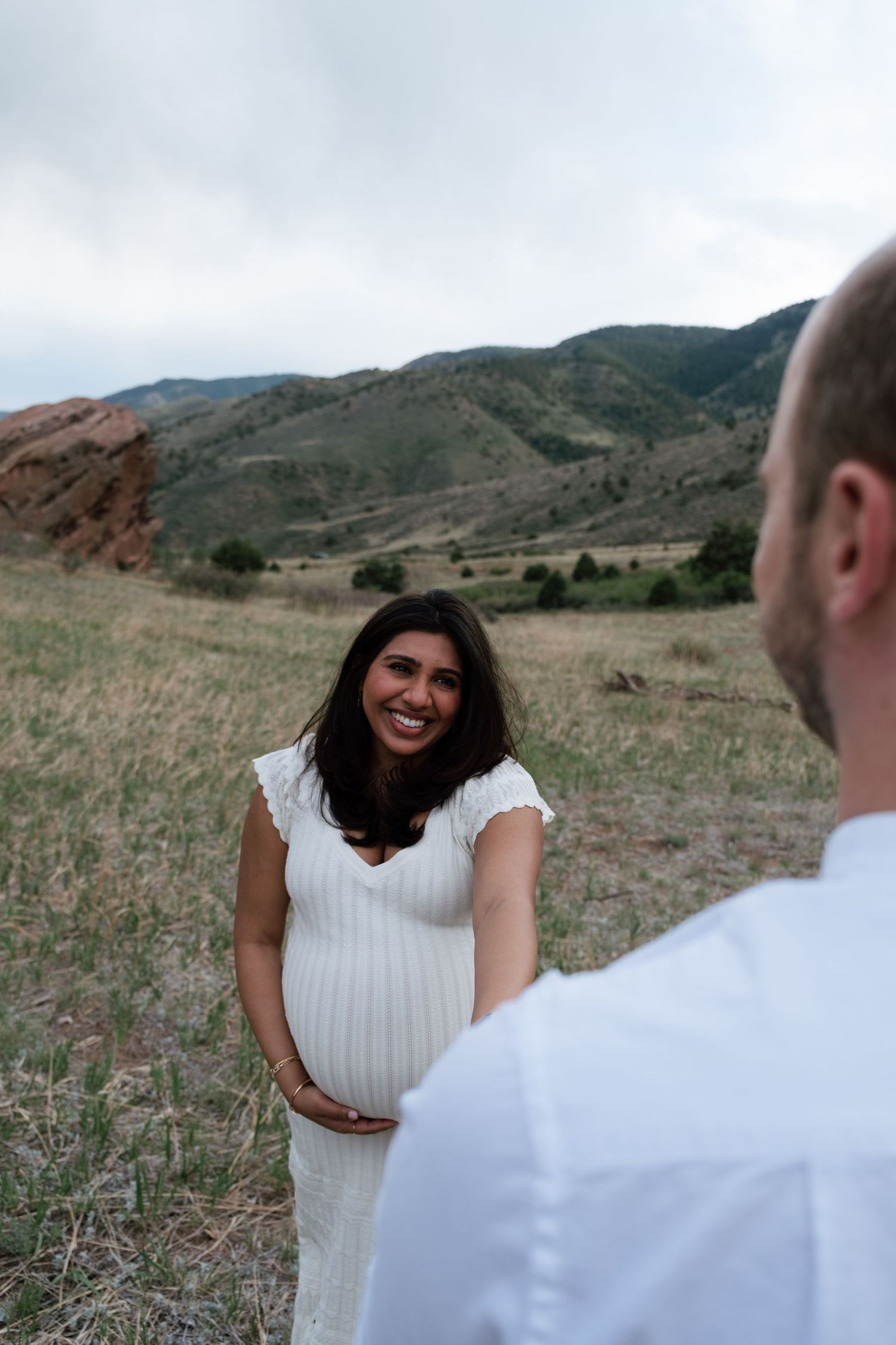 Pregnant woman in white dress smiling at partner in Colorado mountain landscape with red rocks.