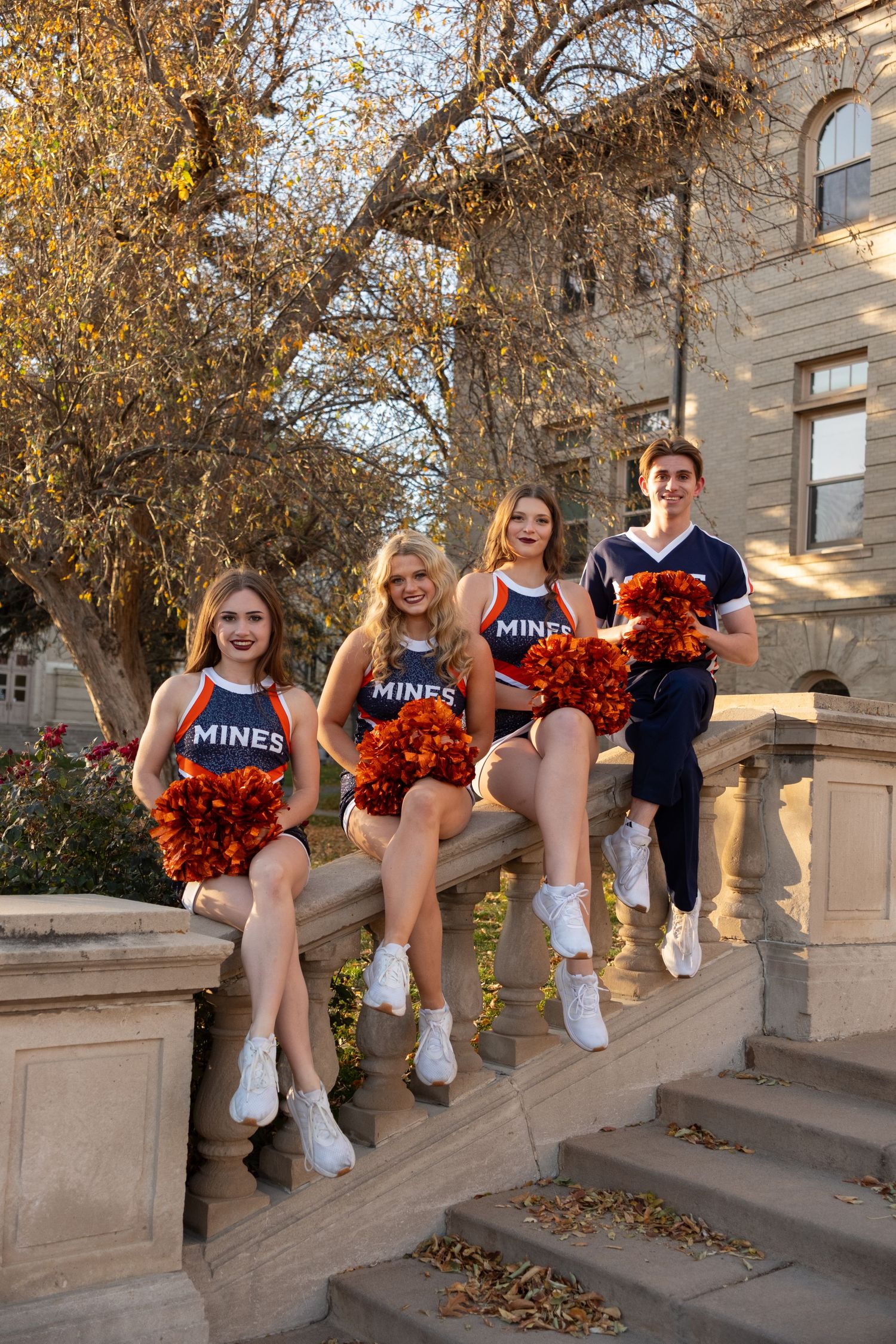 Four Mines cheerleaders sitting on stone staircase railing outdoors, holding orange pom-poms in their uniforms.