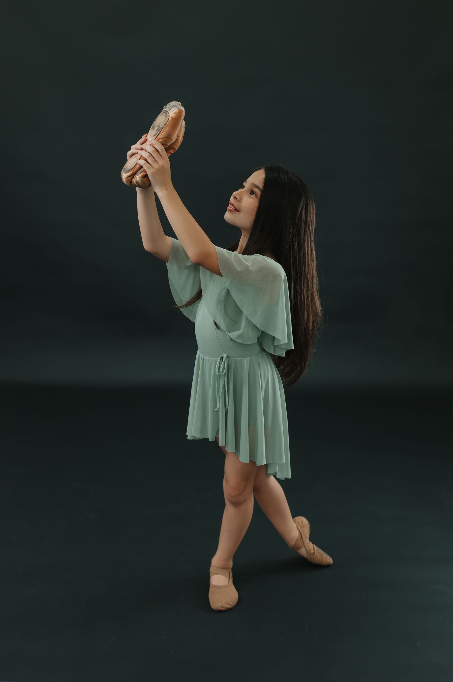 Dancer in mint green dress posing gracefully with pointe shoes against dark background.