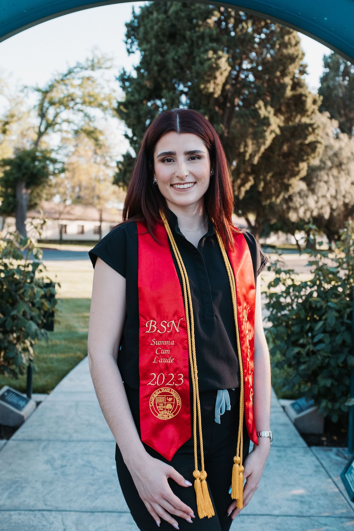 Graduate wearing a black outfit with red graduation stole in an outdoor setting.