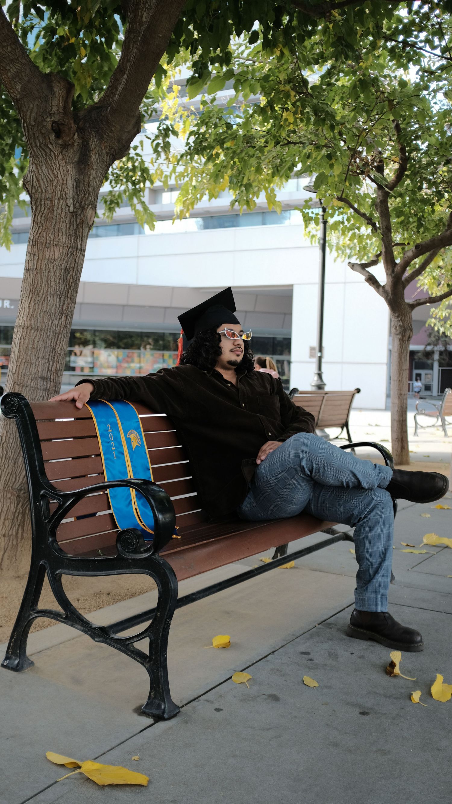 Graduate in cap and gown reclining on a wooden bench with a blue backpack under trees on campus.
