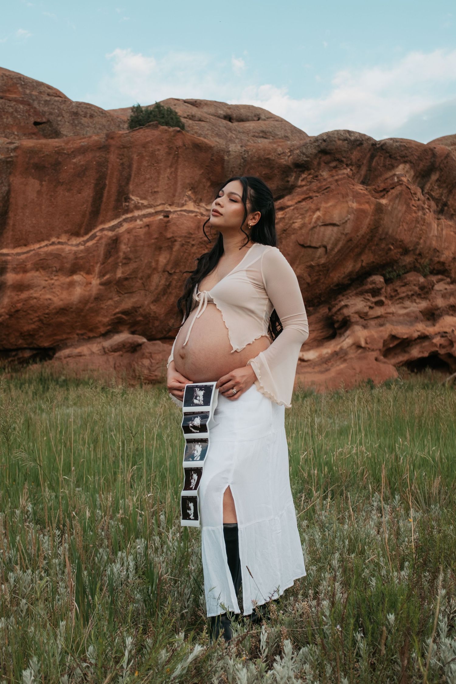 Expectant mother in white dress displays ultrasound photos against dramatic red rock formations.
