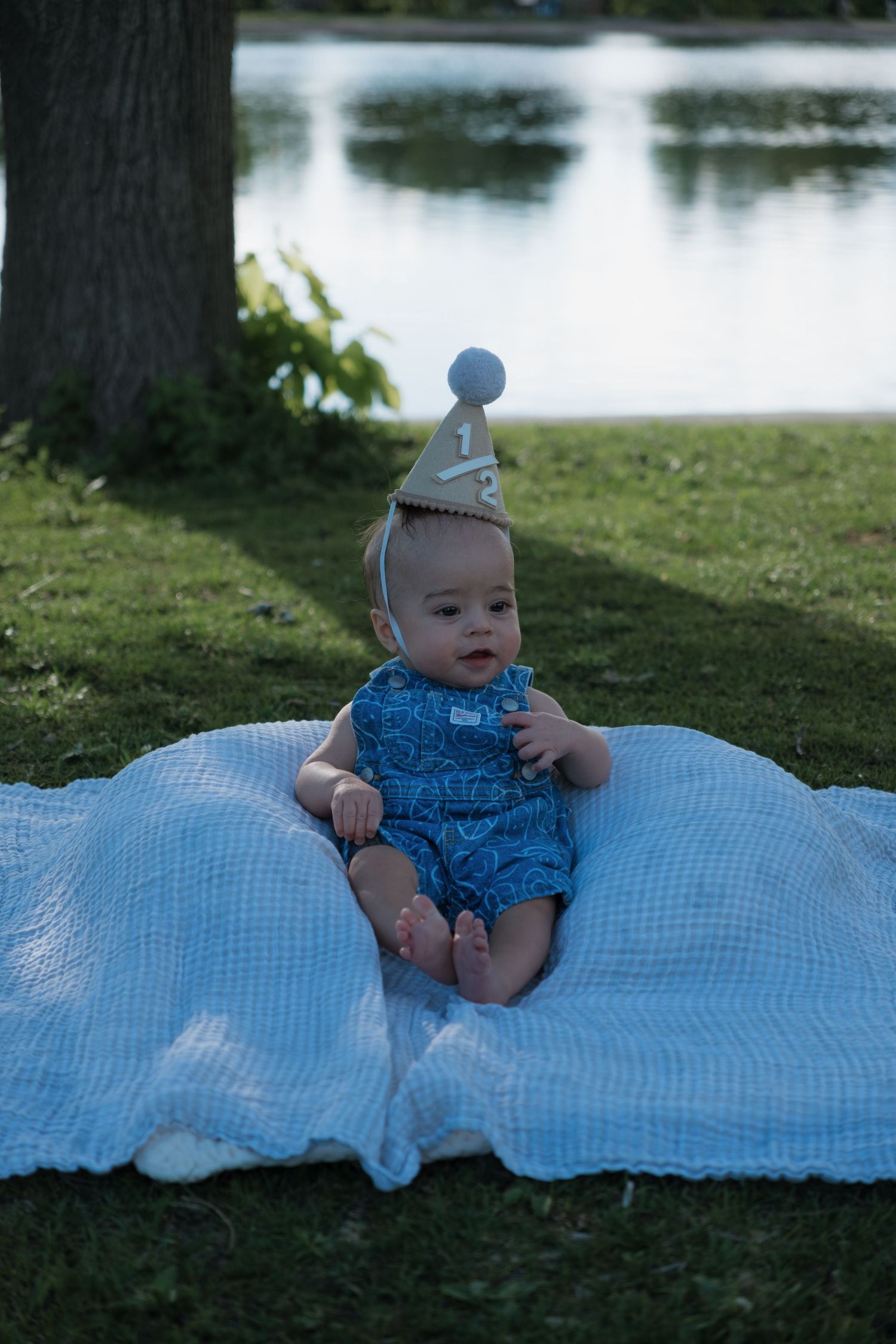 Baby in blue romper and half birthday hat sits on white blanket outdoors near a pond.