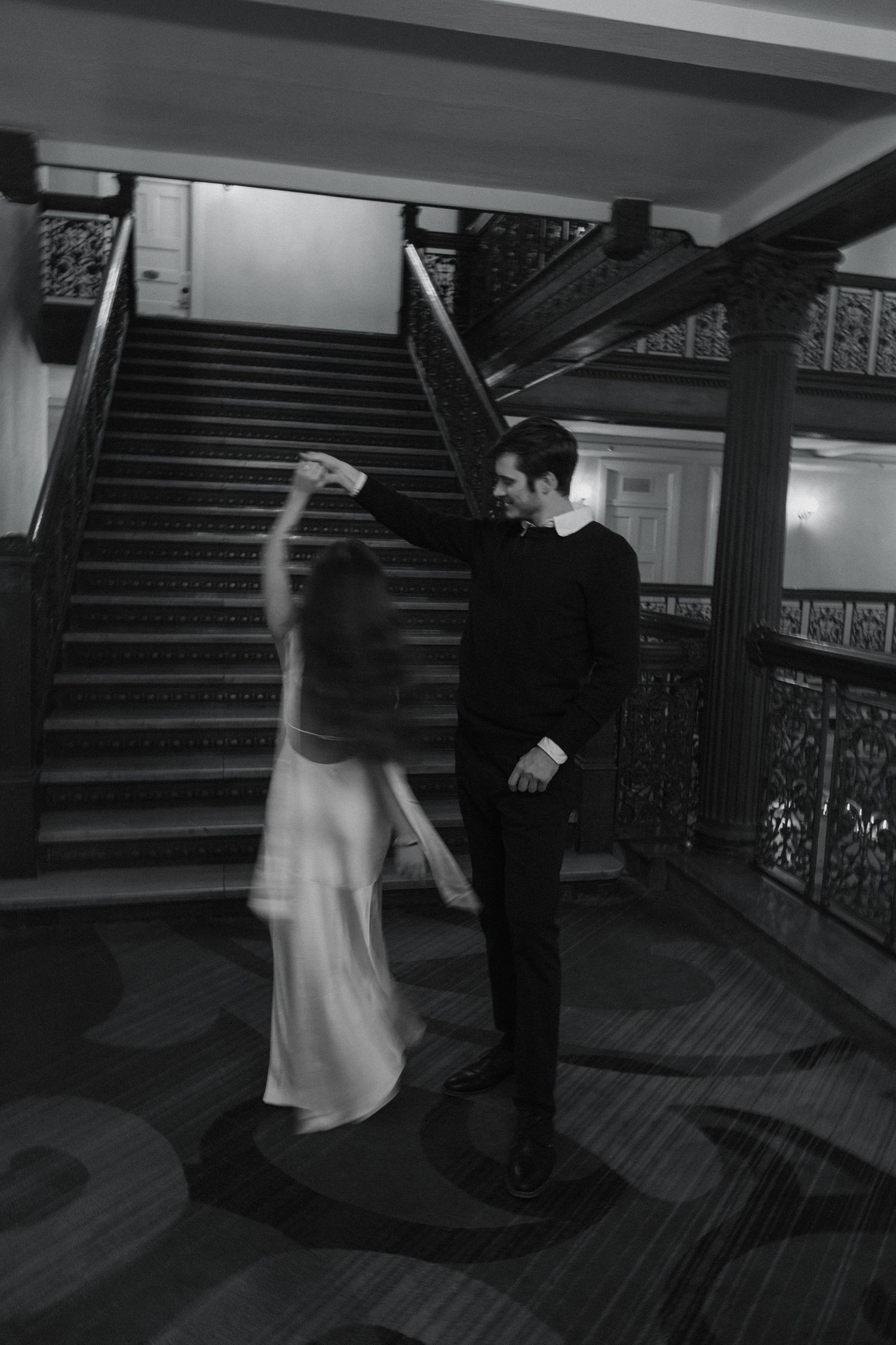 Black and white photo of a couple dancing elegantly near a grand staircase in a historic building.