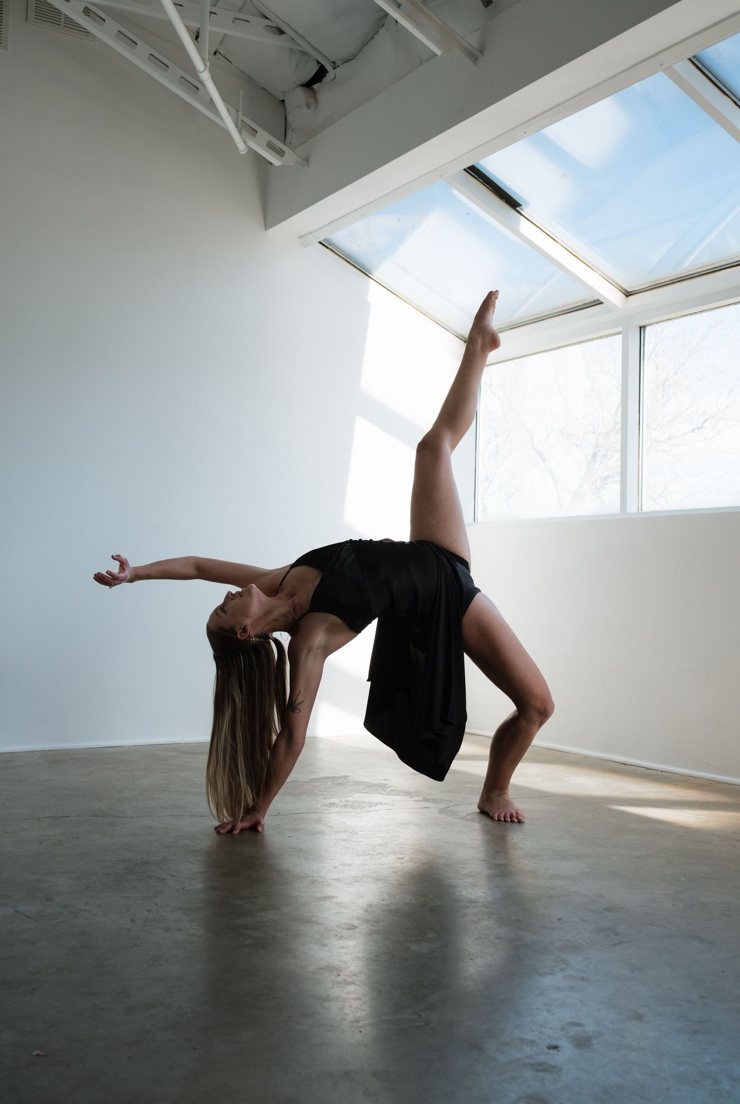 A flexible female dancer in black performs a backbend split on a studio floor with skylights above.
