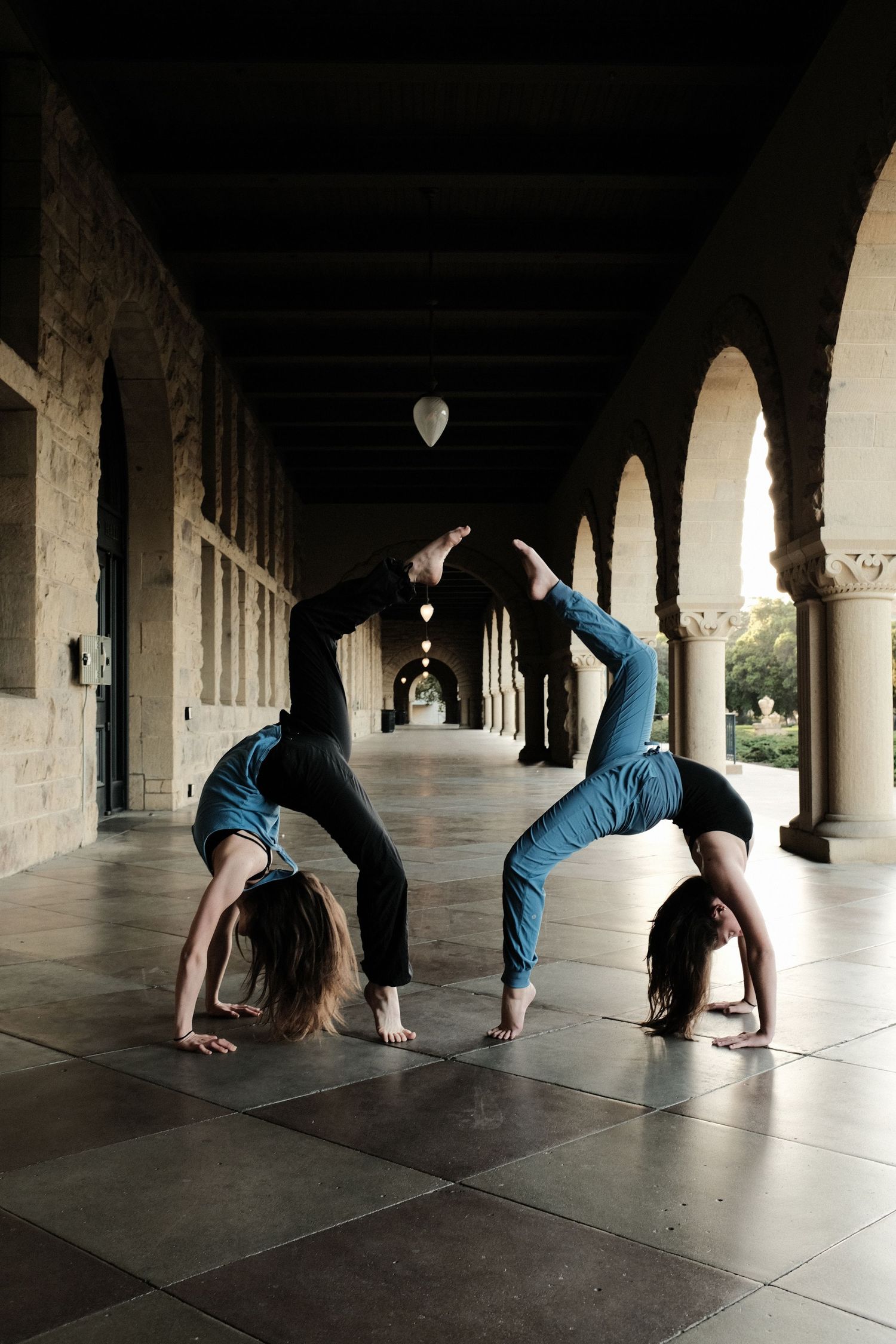 Two dancers perform synchronized back bends in an arched hallway with dramatic lighting.