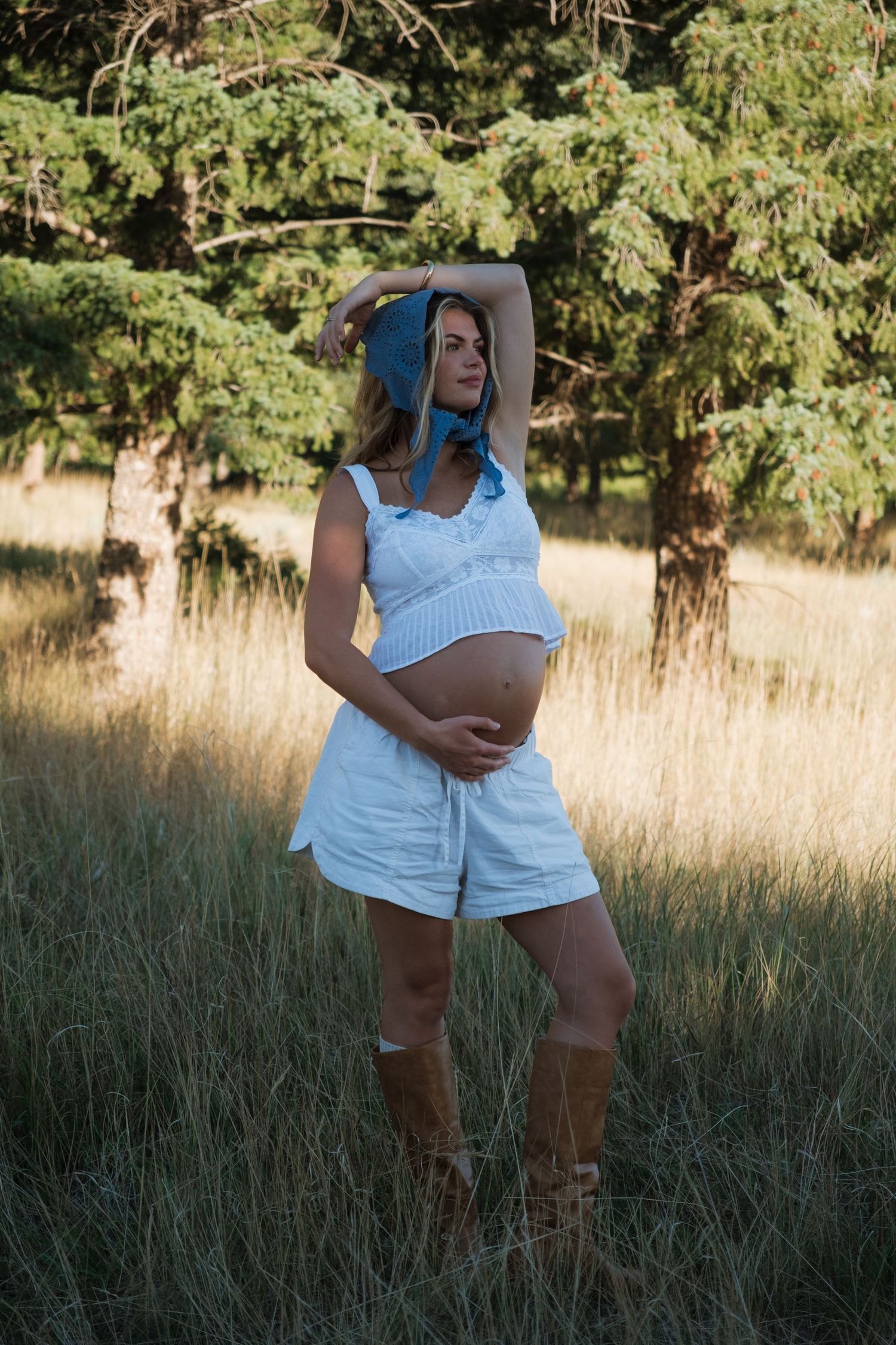 Pregnant woman in a blue headscarf, white crop top and shorts, and brown boots stands in a sunlit grassy field with evergreen trees.