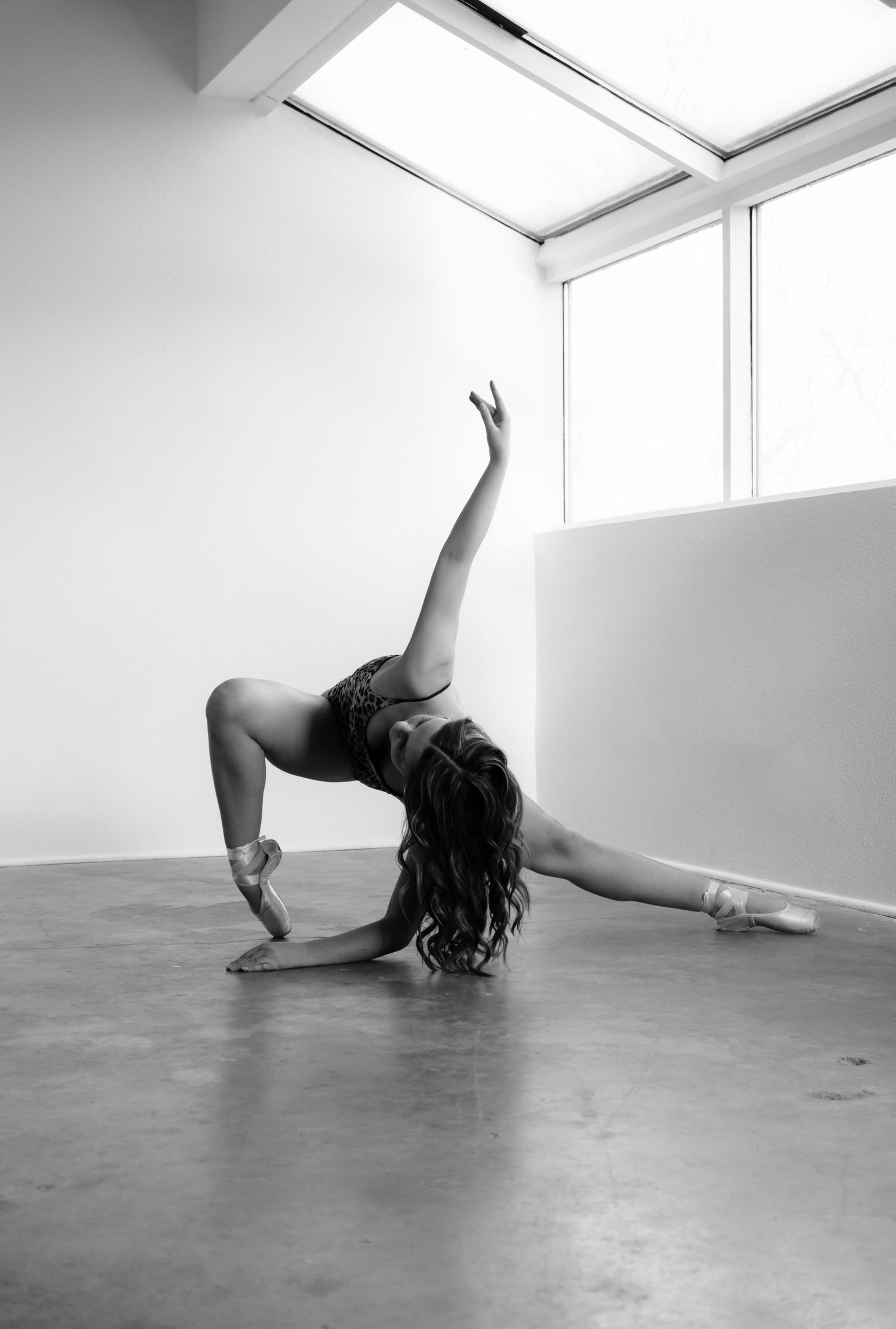 Black and white photo of a ballet dancer in pointe shoes performing an elegant split stretch in a bright studio.