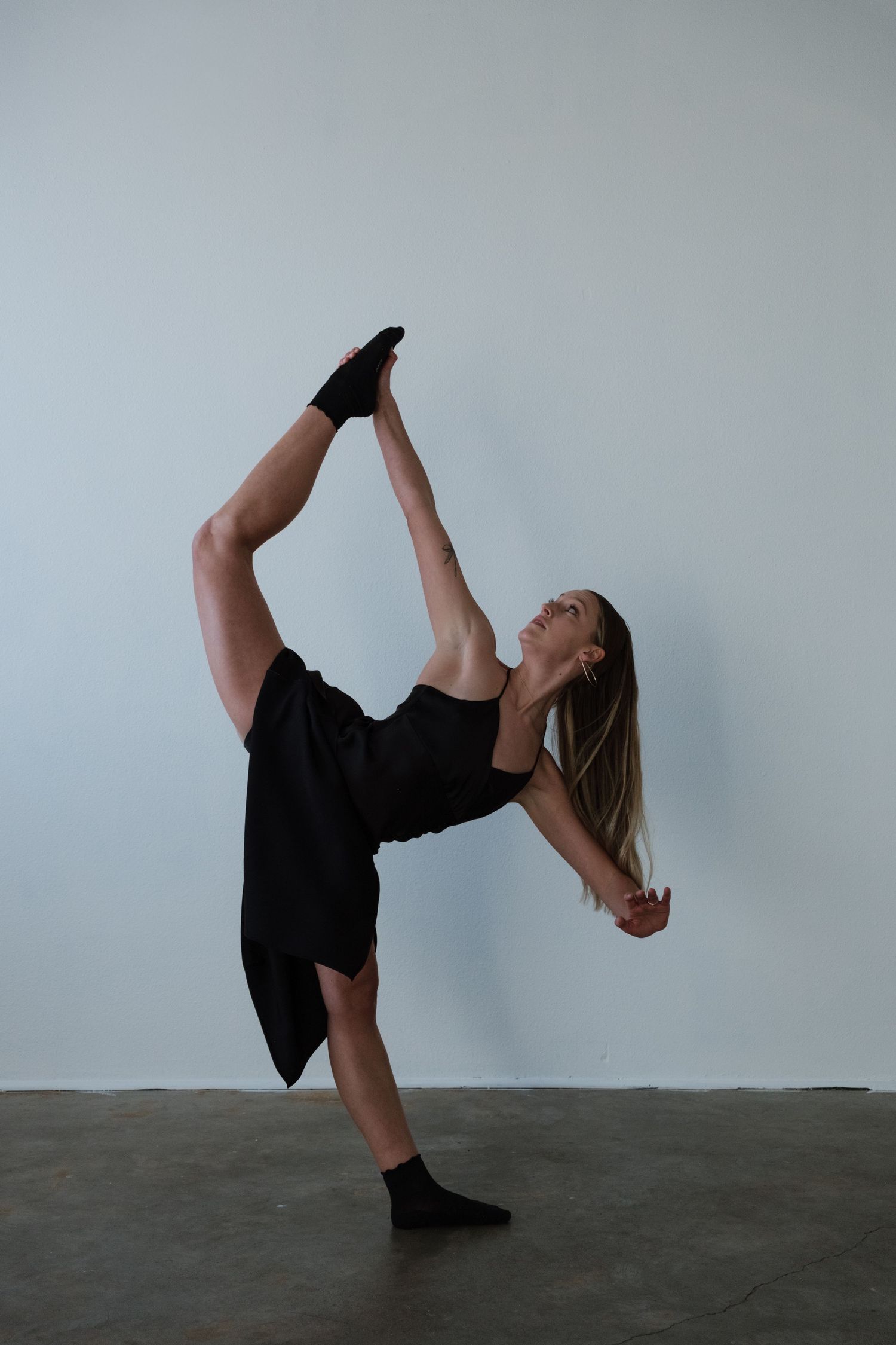 A dancer in black performs a standing split, balancing on one leg with arm extended in a studio.