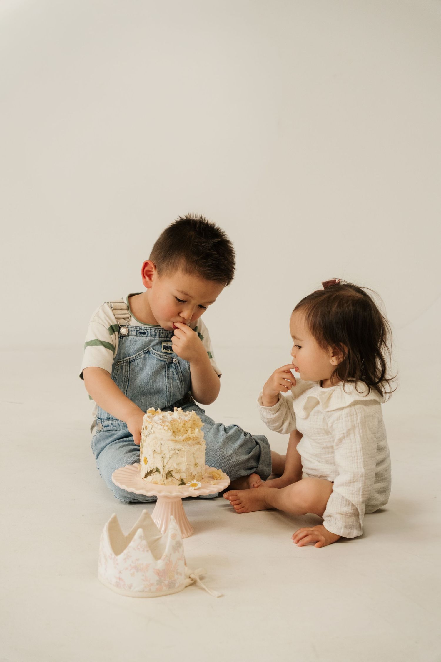 Two young children sitting on floor sharing a smashed white birthday cake during a cake smash photo session.