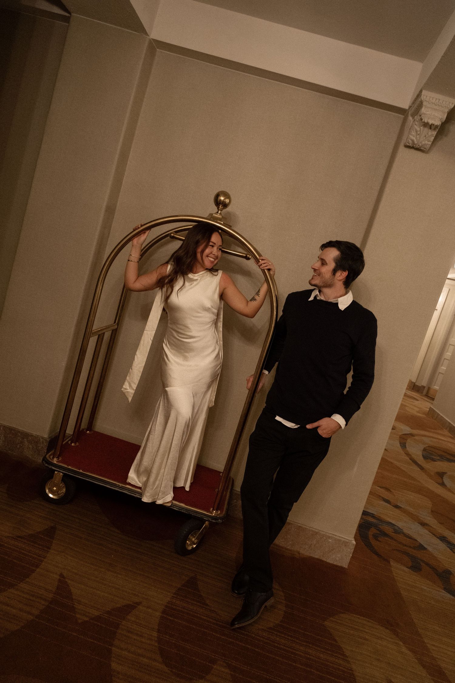 Bride in white gown stands on gold hotel luggage cart while groom in black smiles beside her in hotel hallway.