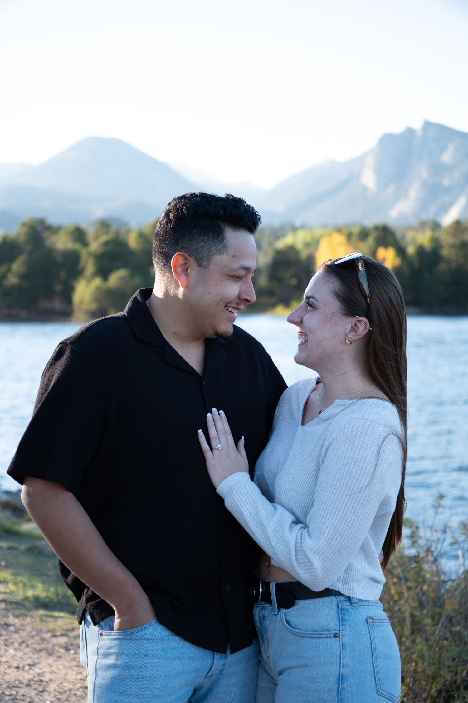 Couple smiling at each other near a mountain lake, woman in white sweater placing hand on man in black shirt's chest.