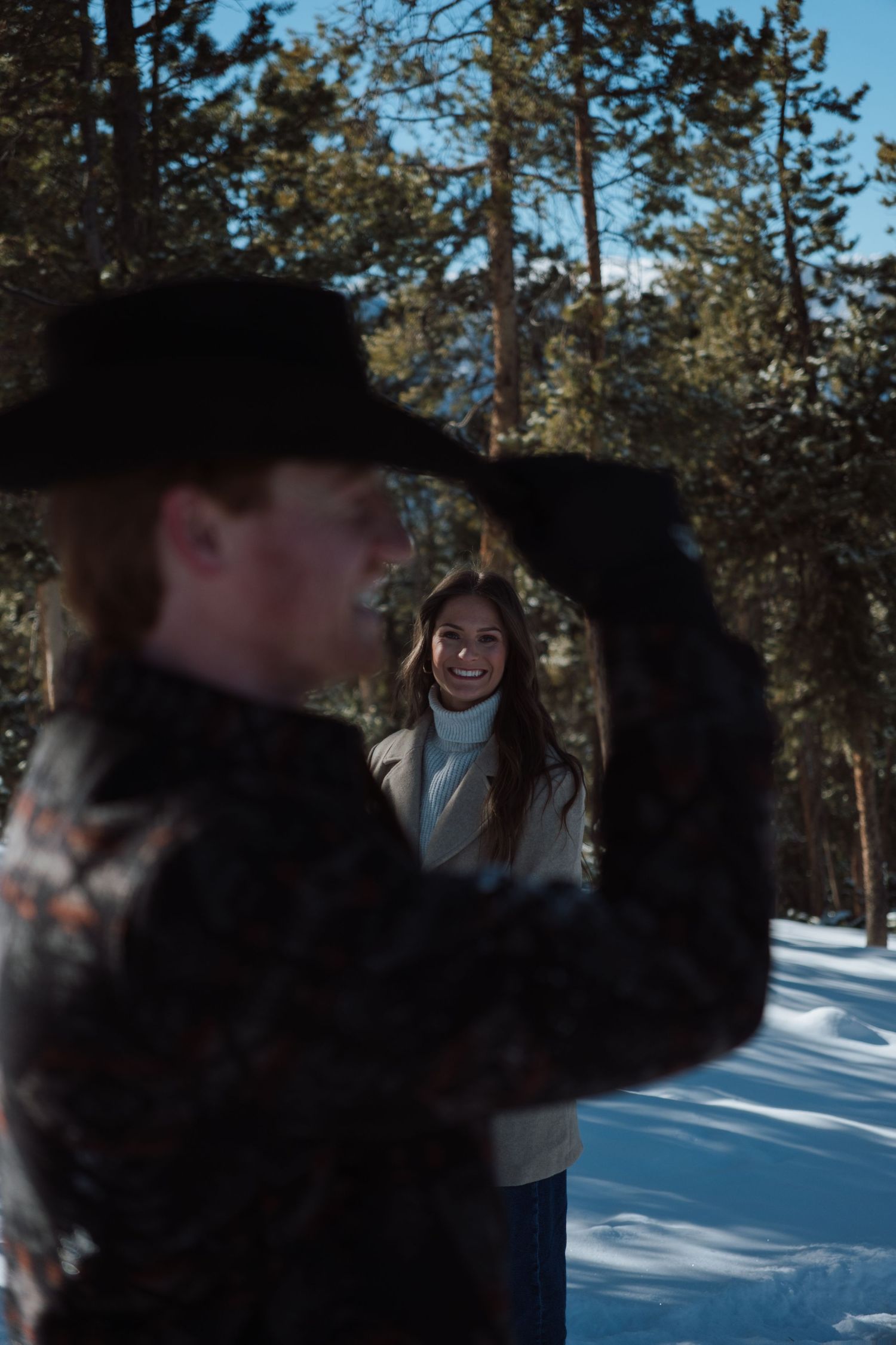Couple in snowy forest, woman smiling in white jacket, man in black cowboy hat in foreground, sunny winter day.