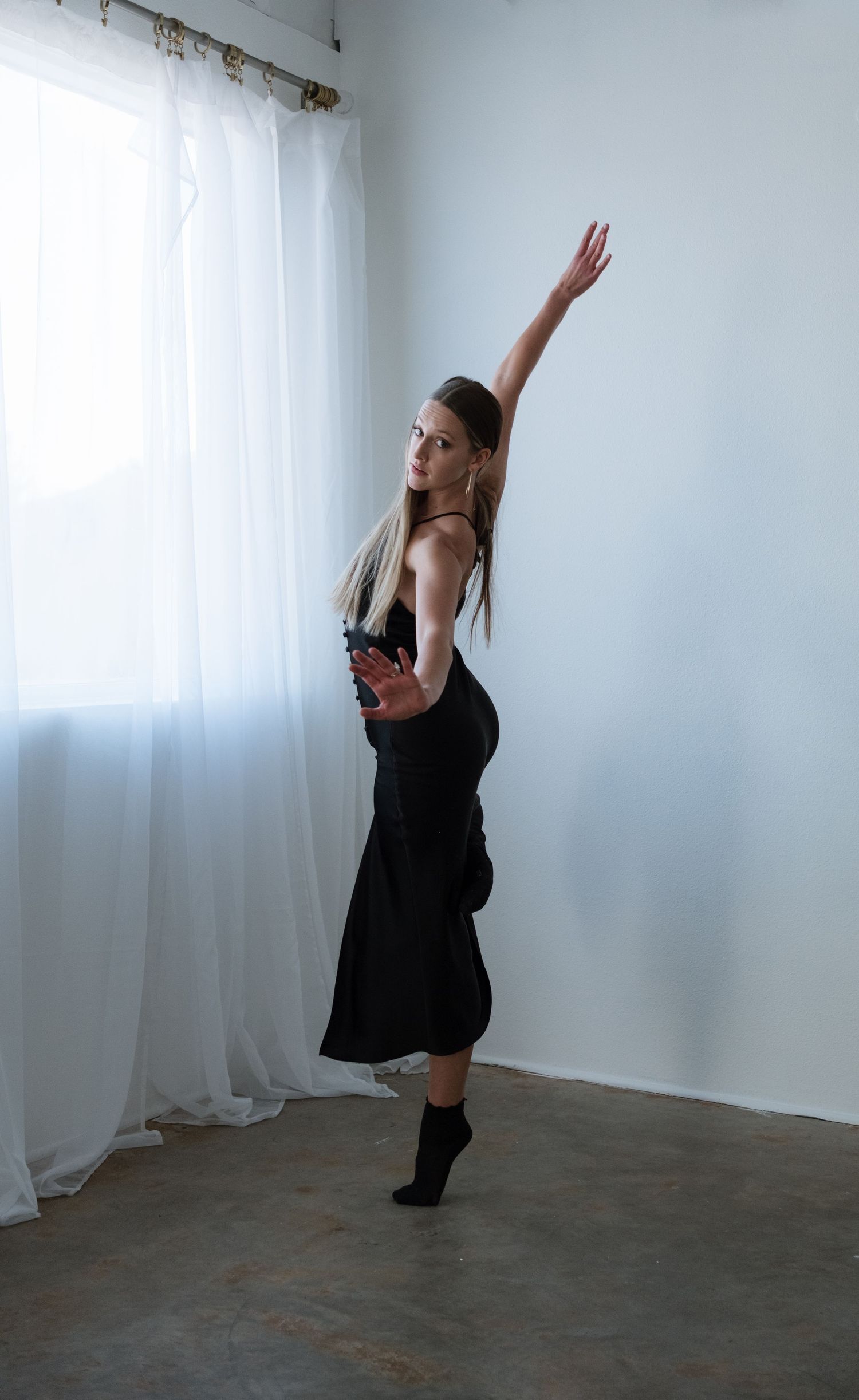 A female dancer in a black dress elegantly poses with one arm raised in a bright studio with white curtains.