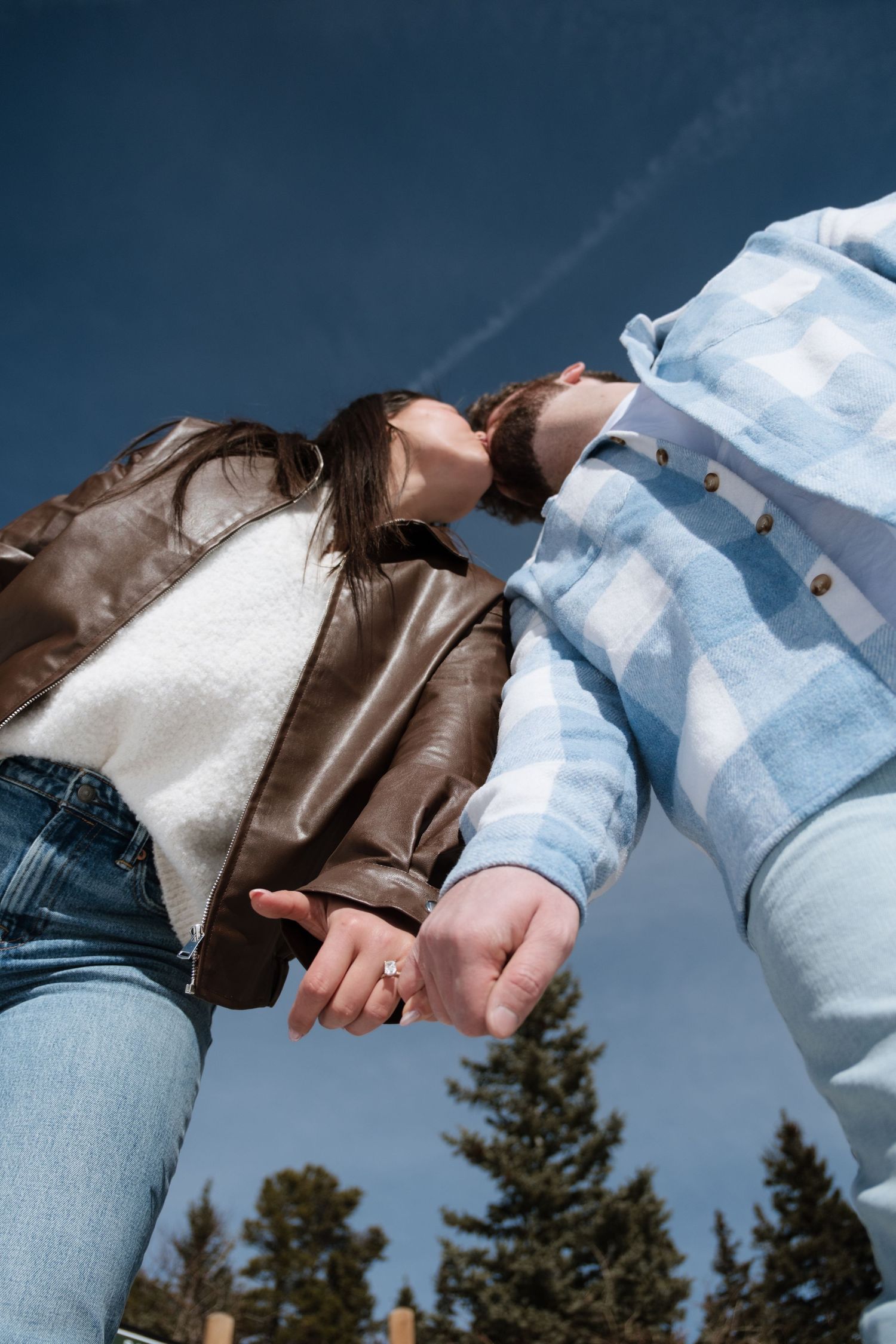 Couple holding hands outdoors under blue sky, woman in brown leather jacket and man in light blue plaid shirt.