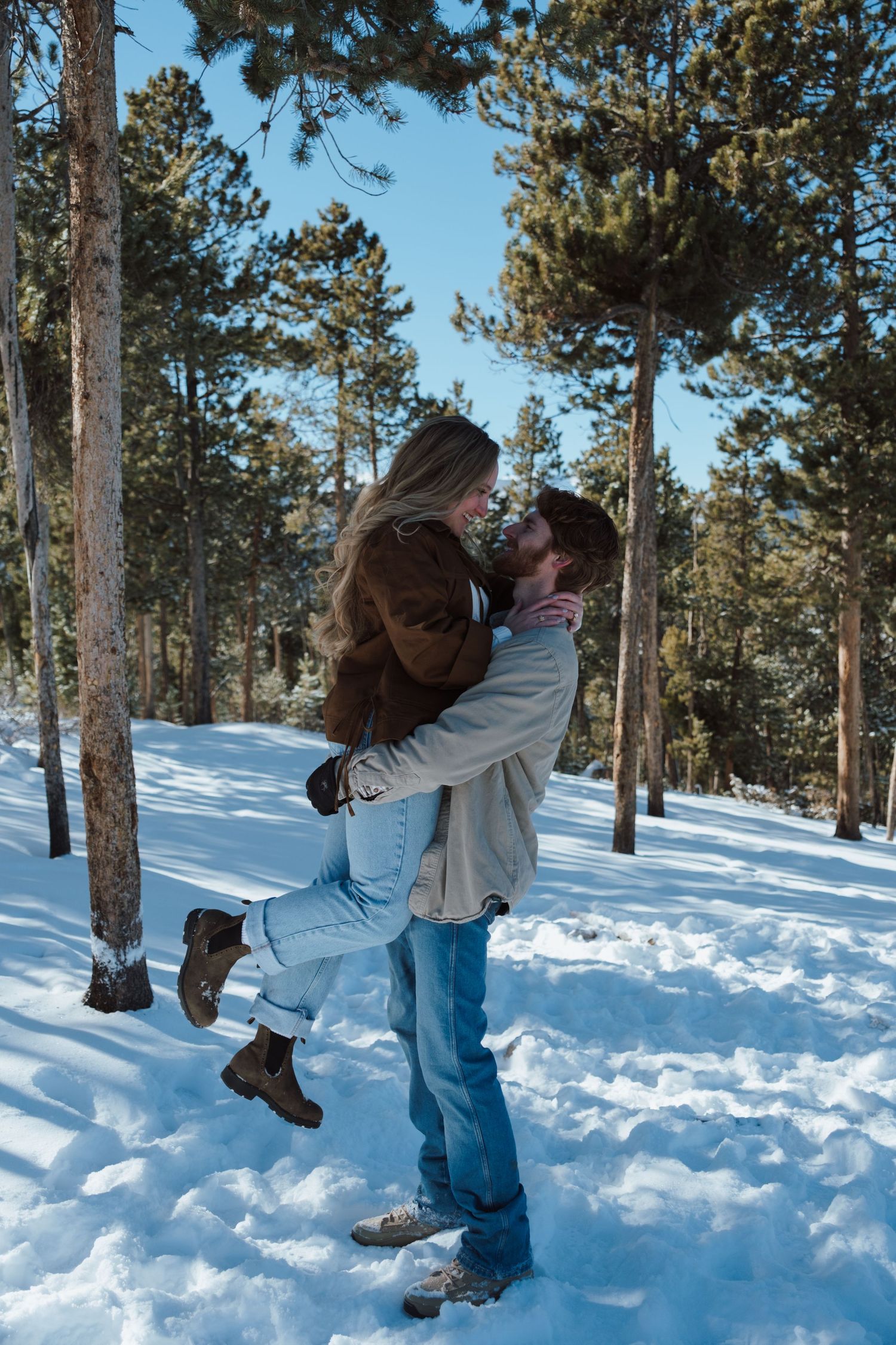 Couple sharing a romantic kiss in a snowy winter forest, man lifting woman in his arms among tall pine trees.