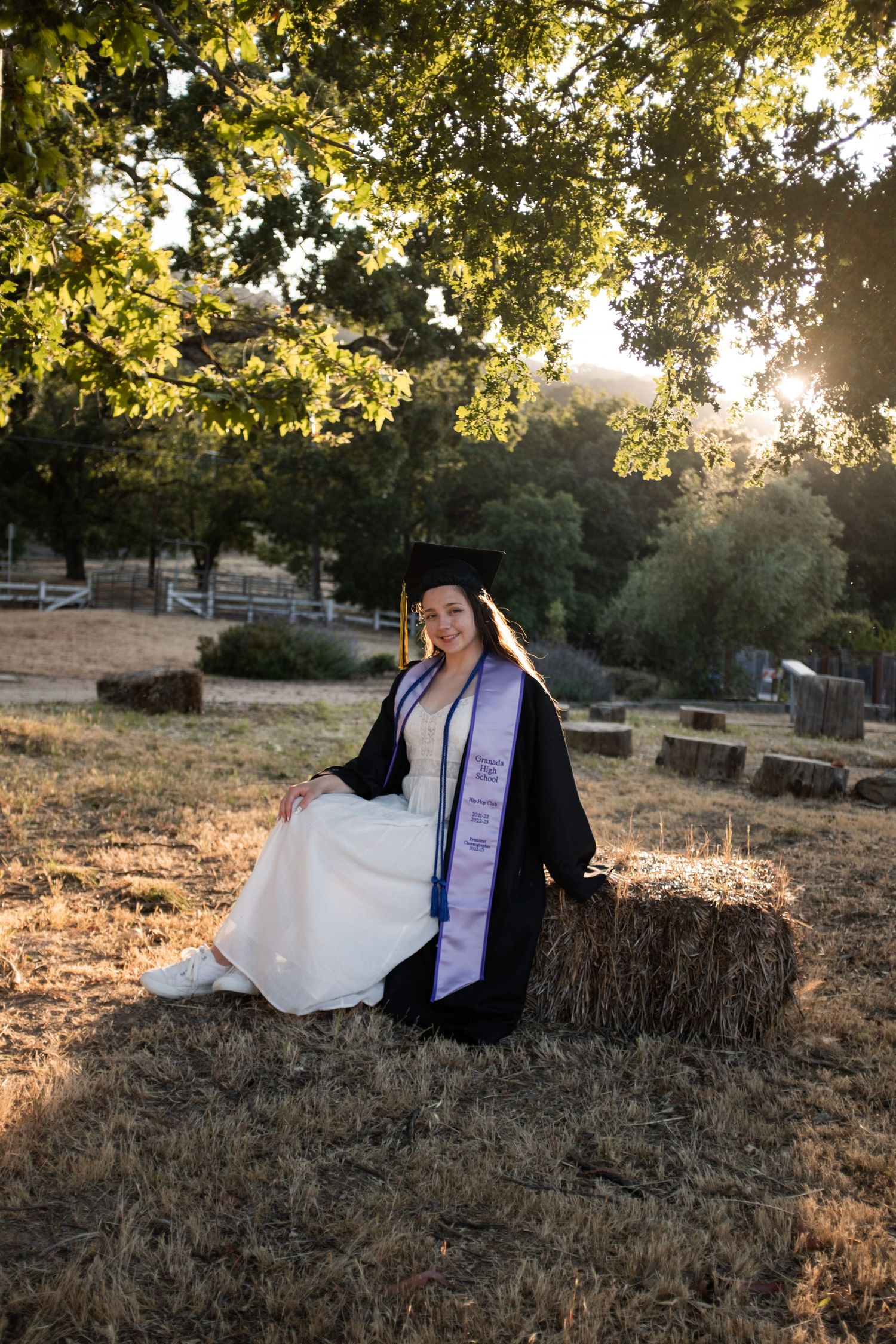 Graduate in cap and gown sits on hay bale during sunset photoshoot.