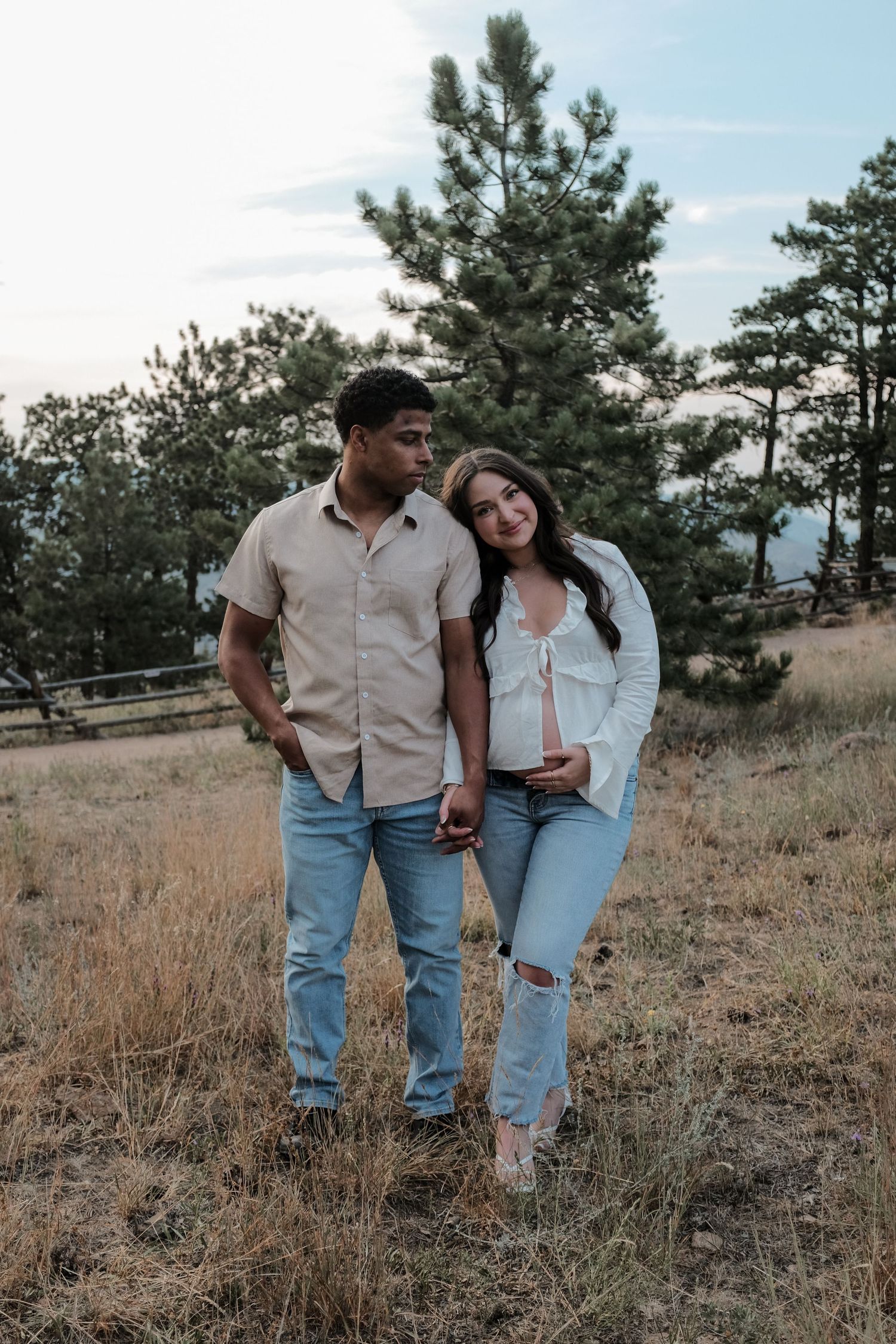 Couple standing in a field with pine trees, woman in white top holding baby bump, man in beige shirt looking at her lovingly.