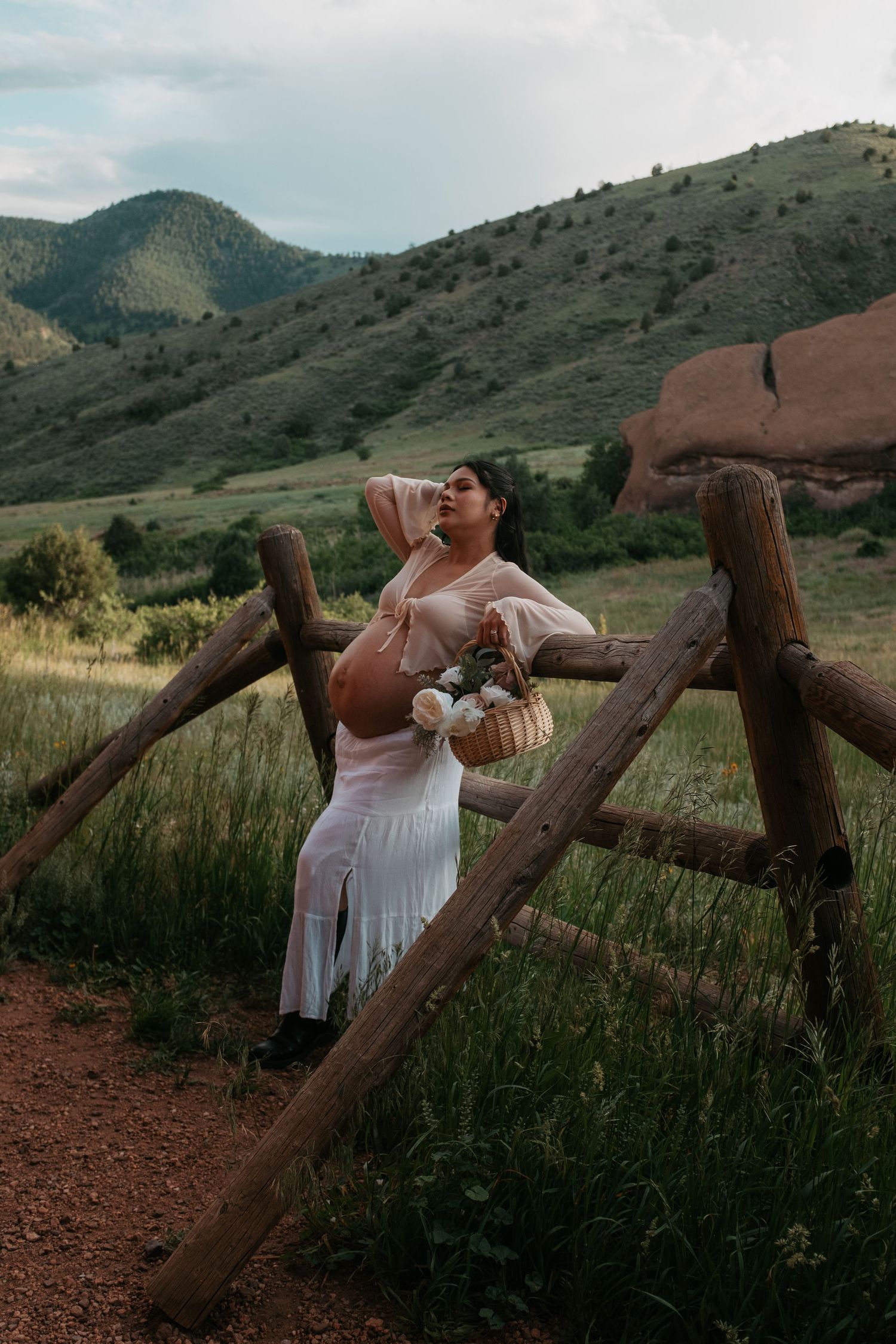 Pregnant woman in white dress poses by rustic wooden fence with mountain landscape and green hills in background.