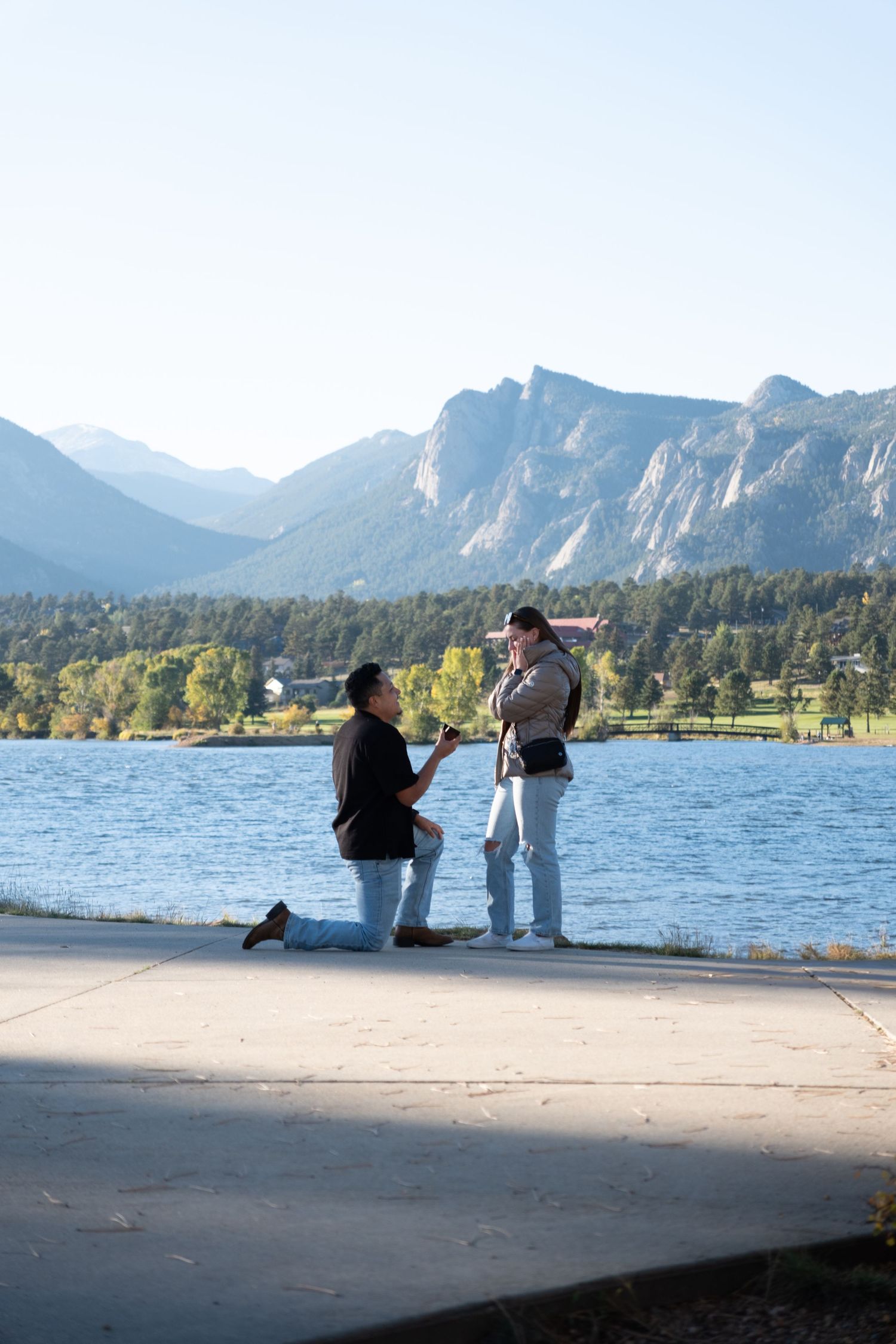 Person proposing on one knee by a mountain lake with stunning Rocky Mountain scenery in the background.