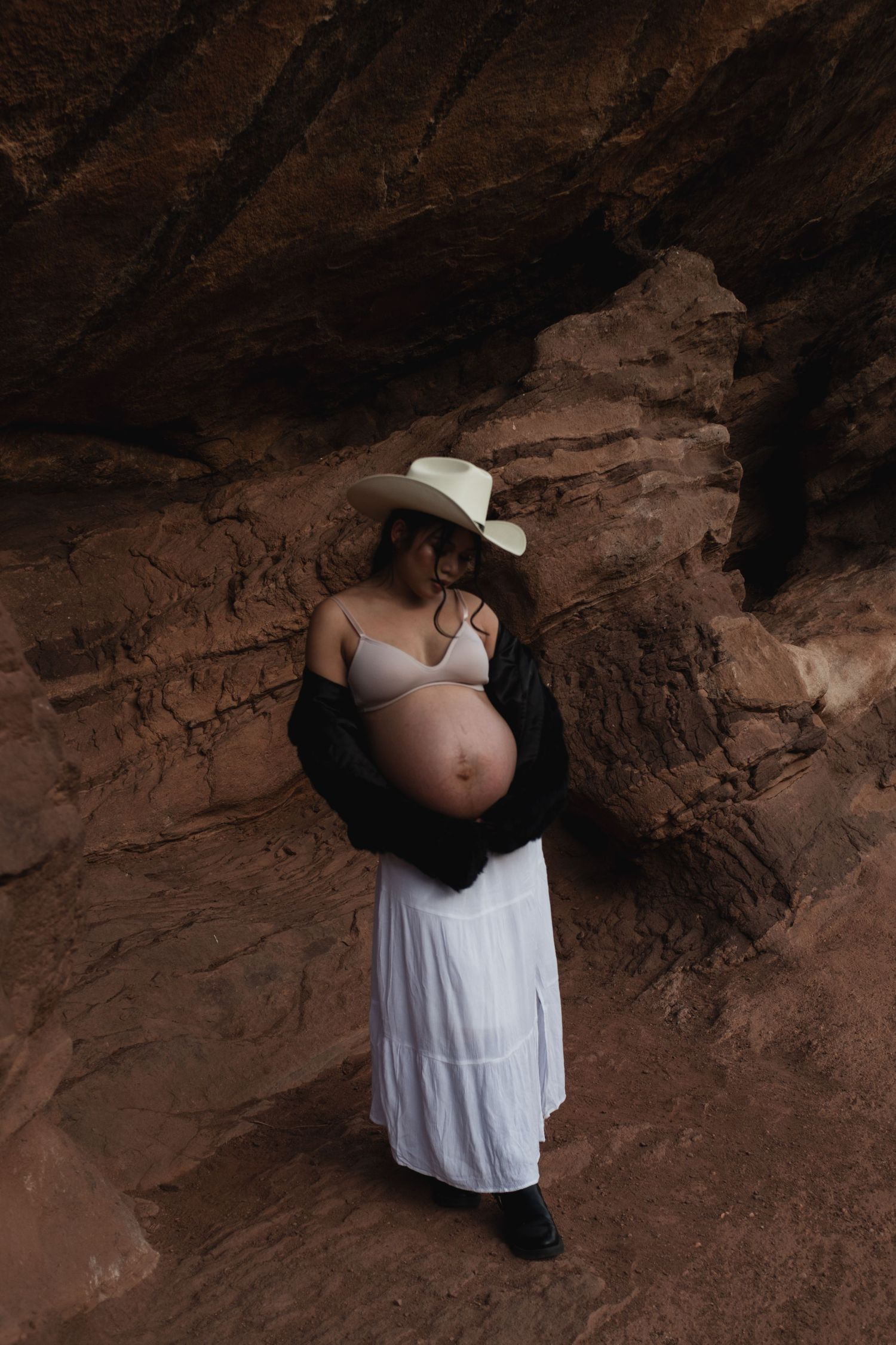A maternity photo shoot in a red rock cave features a woman in a white dress and black cardigan.