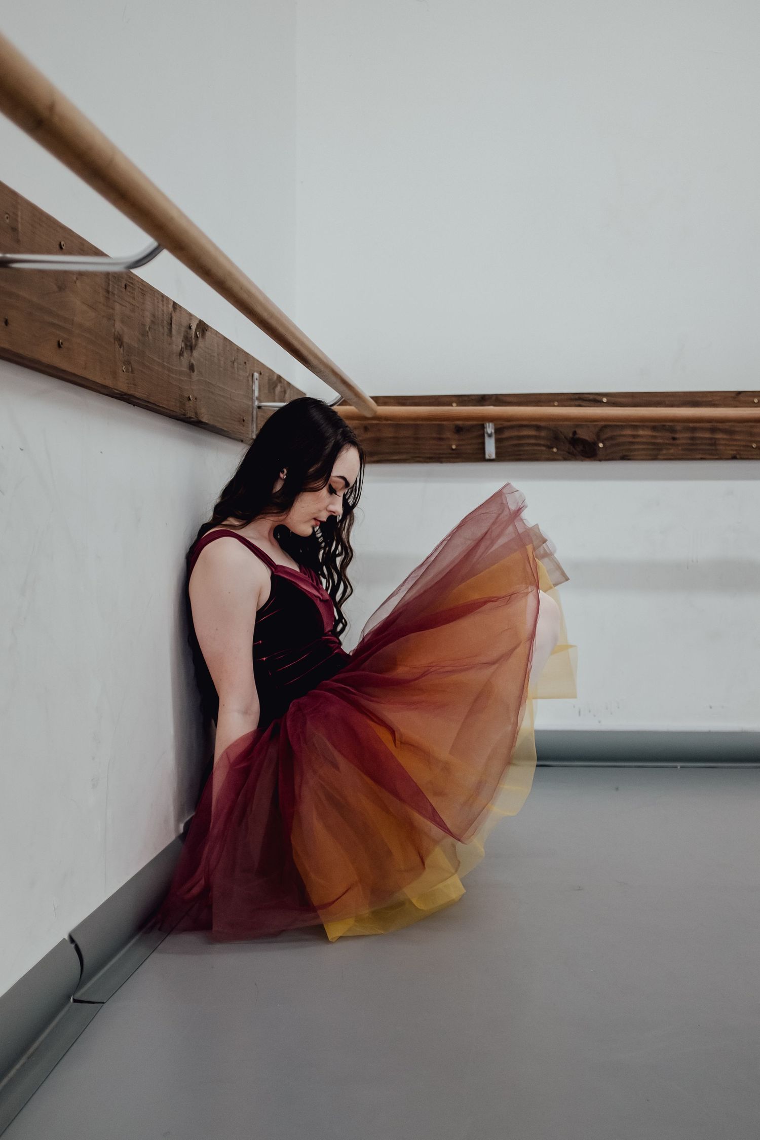 Woman in burgundy tulle dress sitting against white wall in artistic pose.