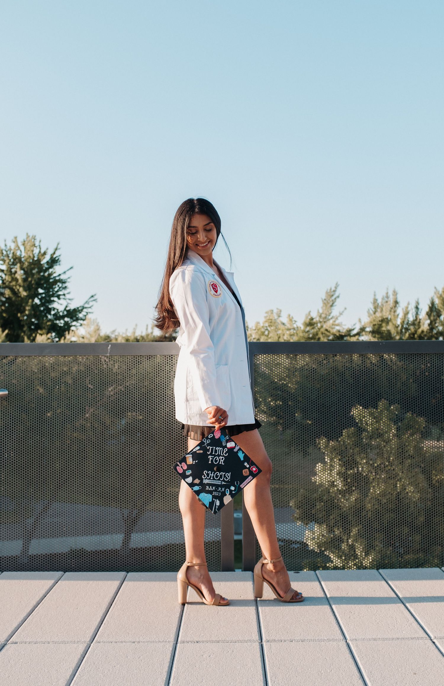 Graduate in white dress posing with decorated cap on outdoor terrace.