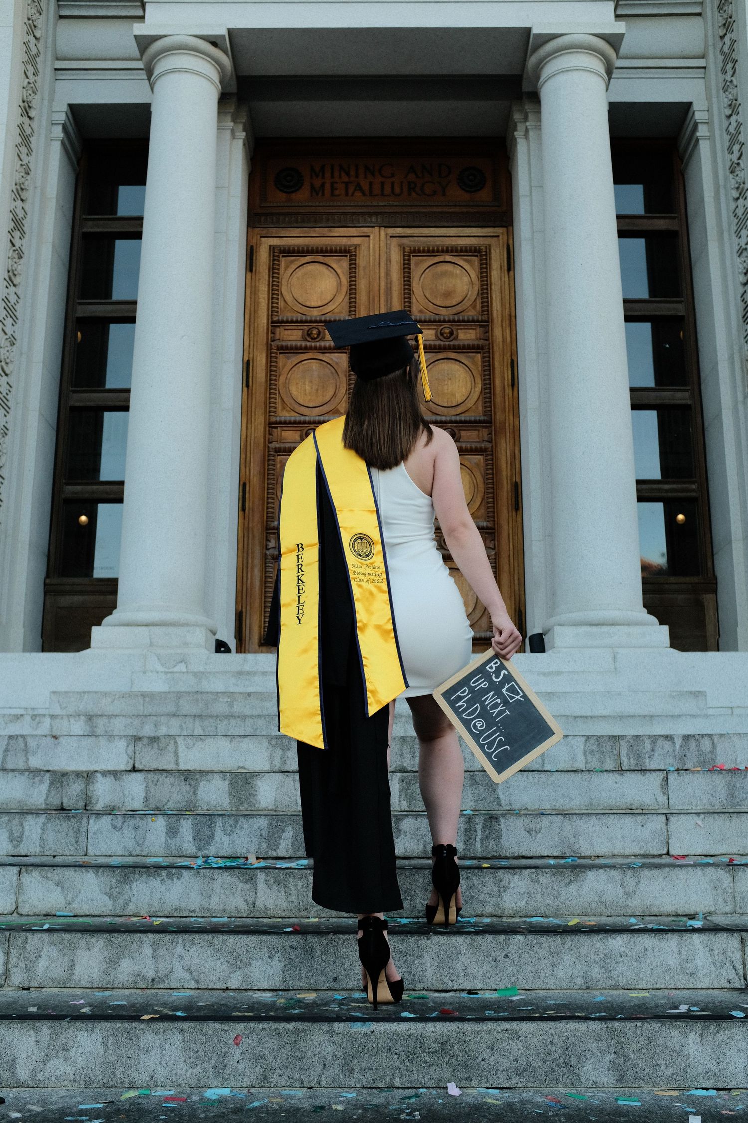 Graduate in white dress and yellow sash stands on stone steps in front of ornate wooden doors.