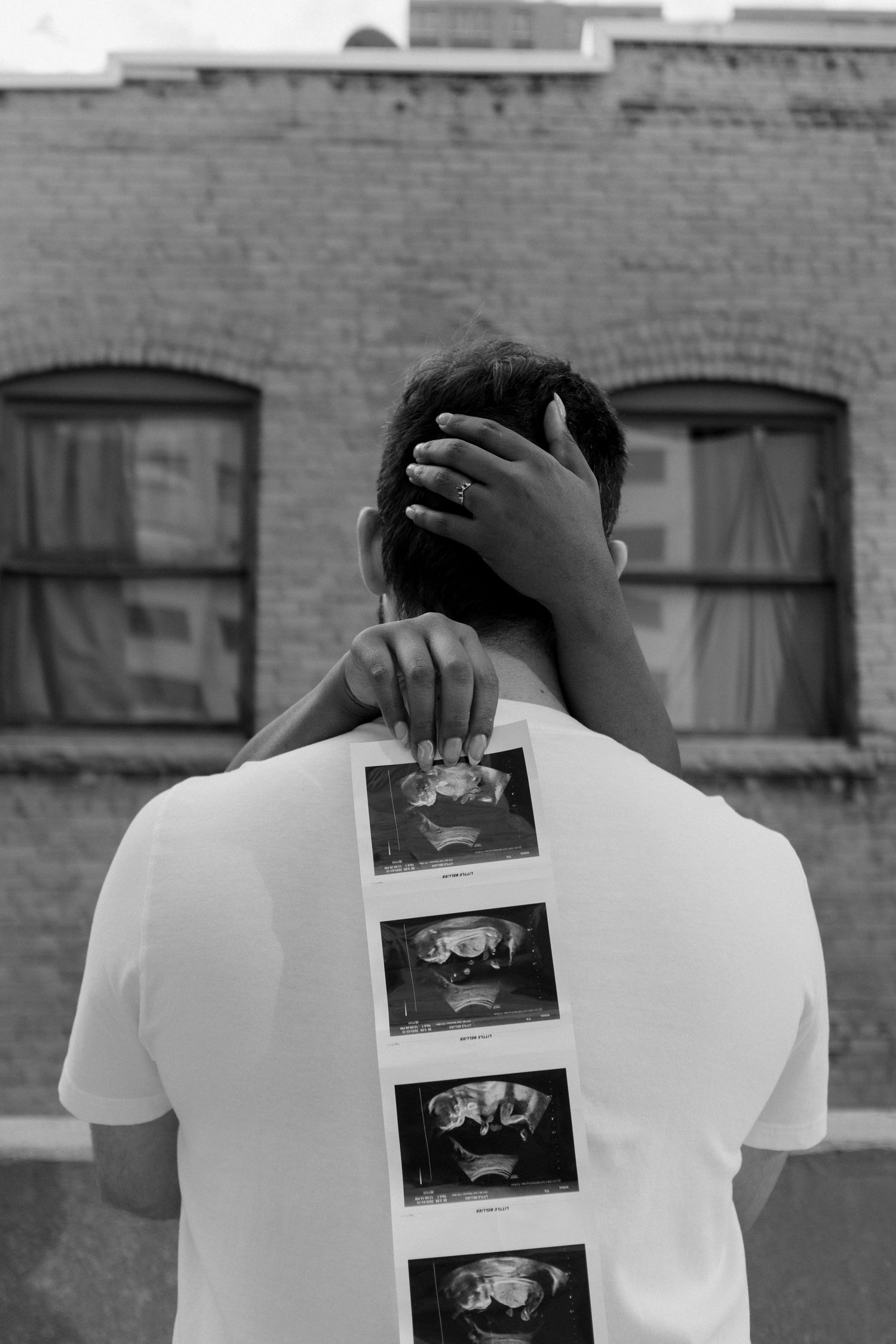 Black and white photo of a person holding ultrasound images on their back against a brick building backdrop.