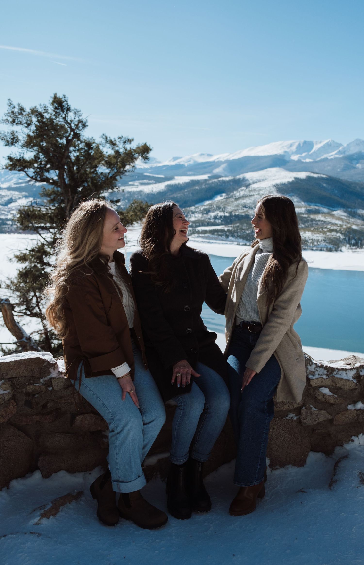 Three women laughing together on a snowy mountain overlook with a frozen lake and peaks in the background.