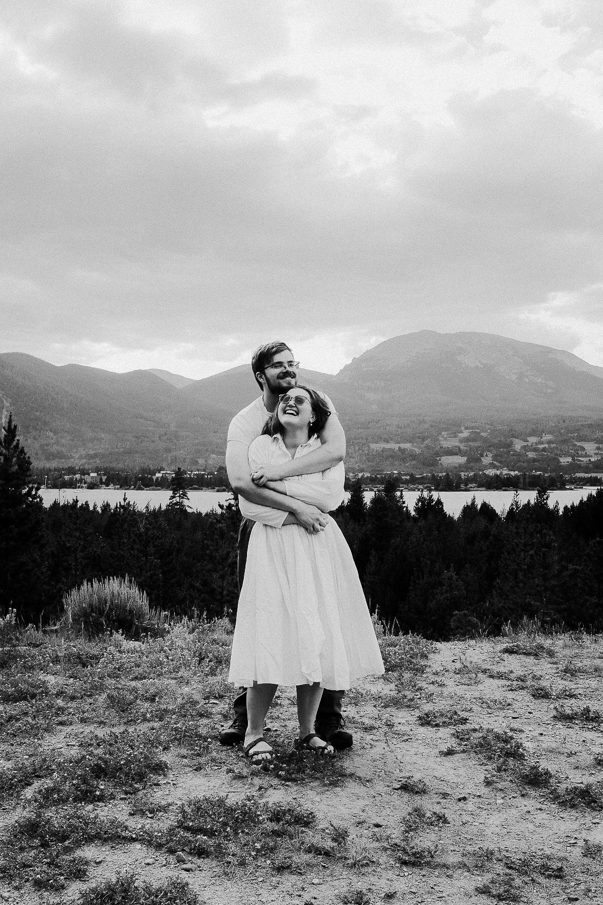 Couple embracing on hilltop overlooking mountain lake landscape in black and white.