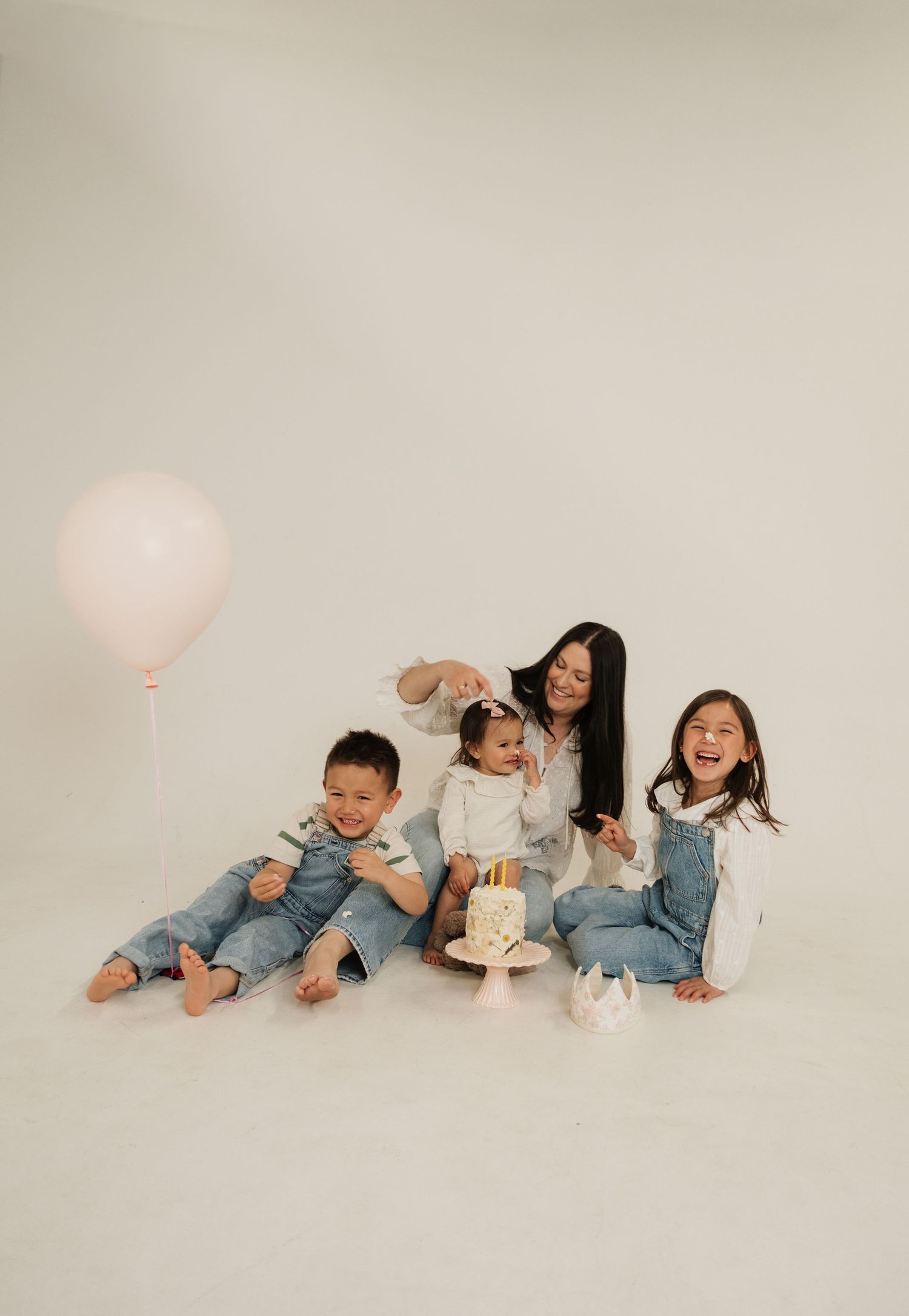 A mother sits on a white studio floor with three young children, a small birthday cake, and a pink balloon.