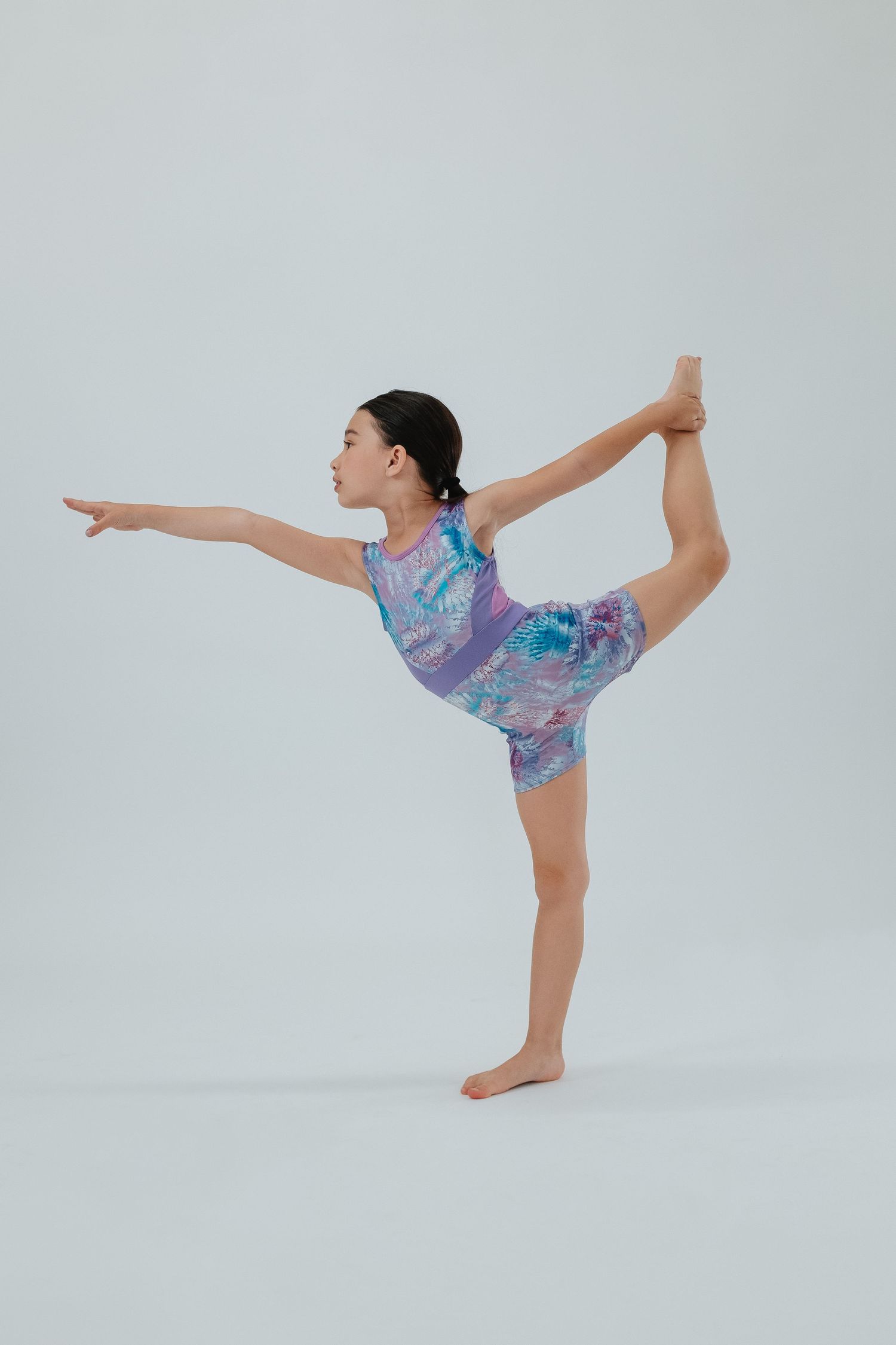Dancer in purple patterned leotard performs standing split pose against white backdrop.