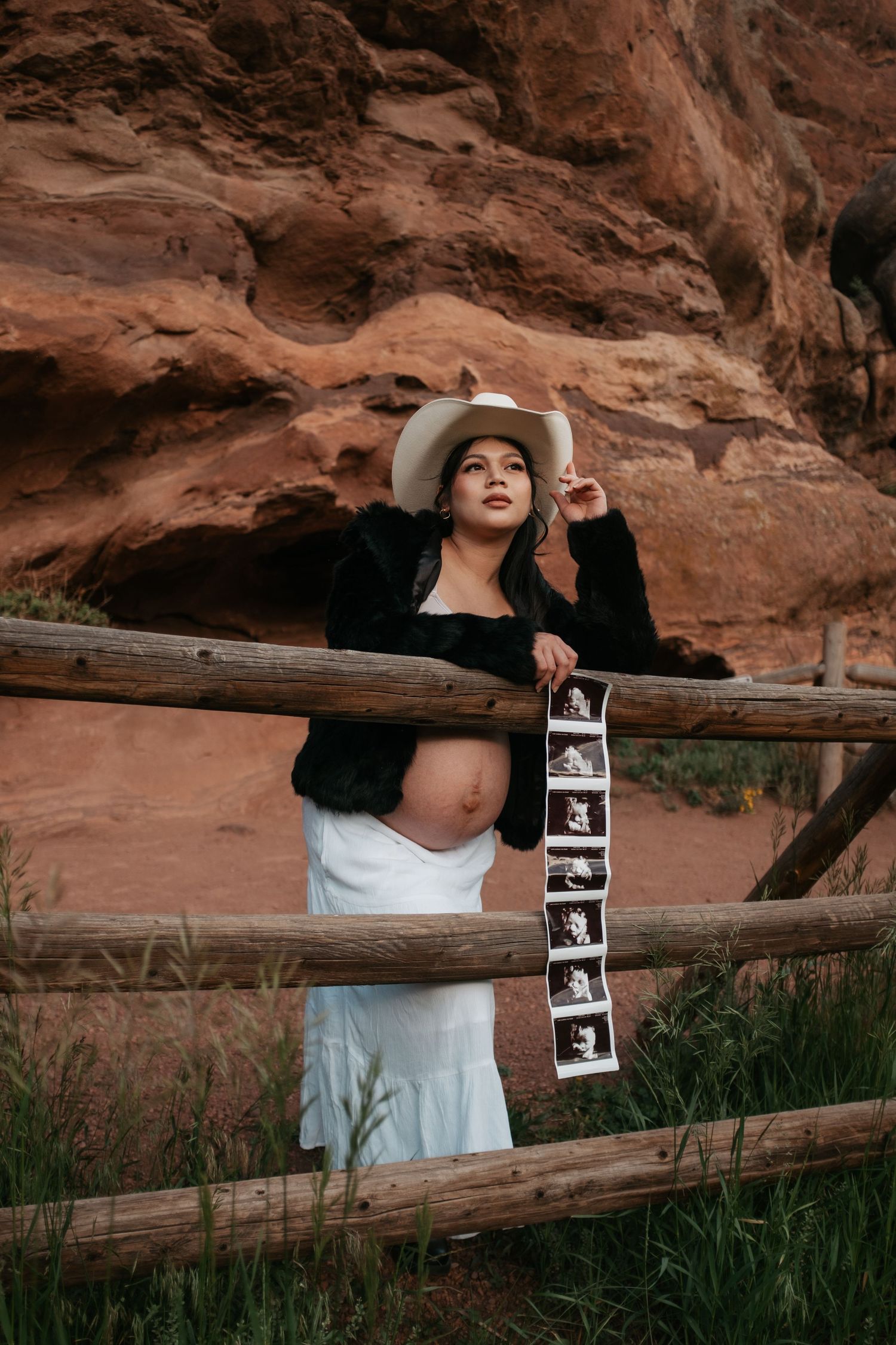 Artistic maternity photo with sonogram strip displayed on wooden fence against red rocks.