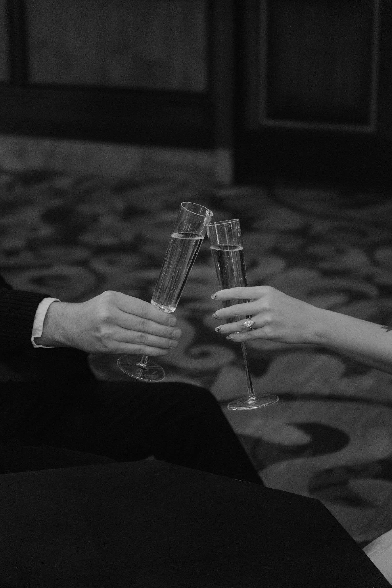 Black and white photo of two hands clinking champagne flutes in a celebratory toast, woman wearing engagement ring.
