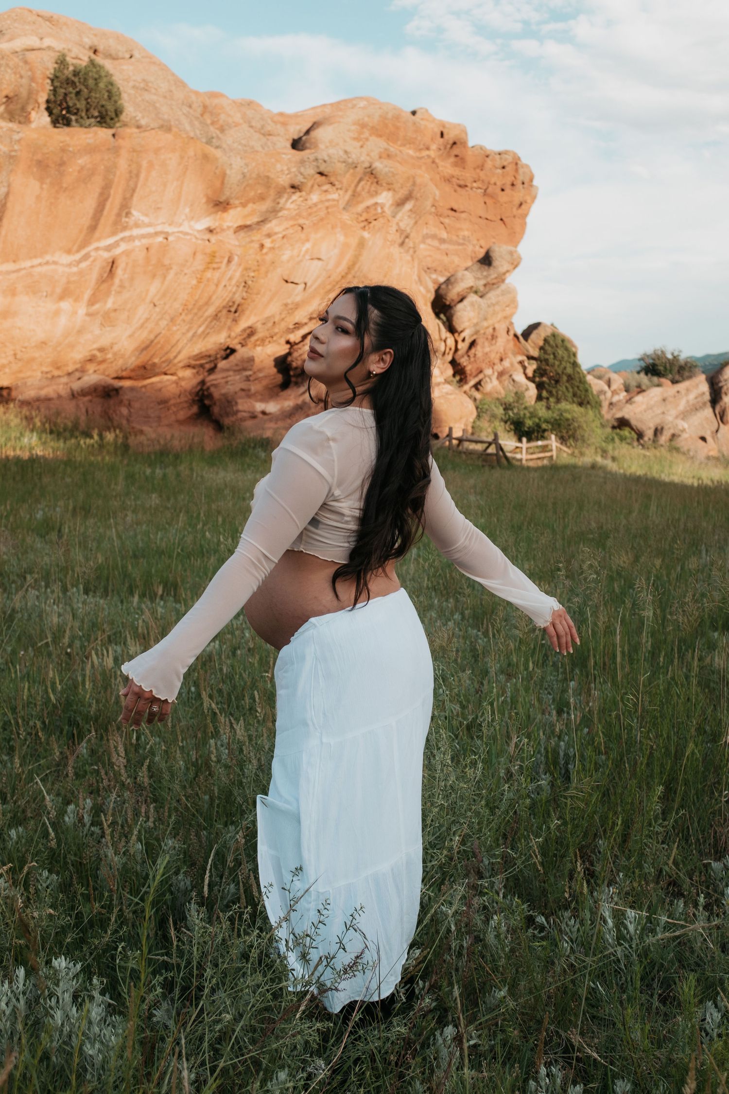 Woman in white maxi skirt and crop top poses in grassy field with red rock formations in background.