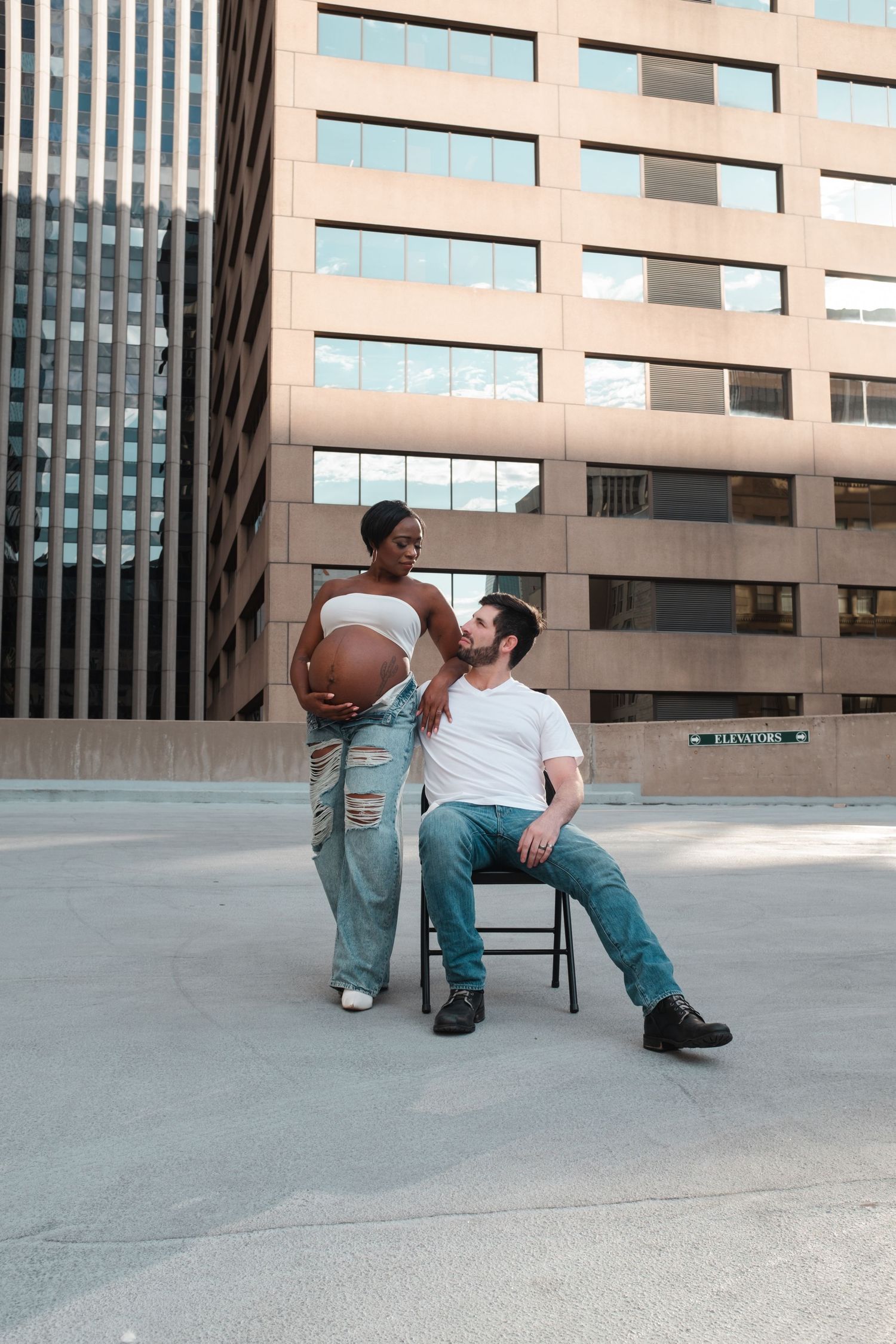 Pregnant woman in jeans stands beside seated man in white tee, sharing a tender moment outdoors near urban buildings.