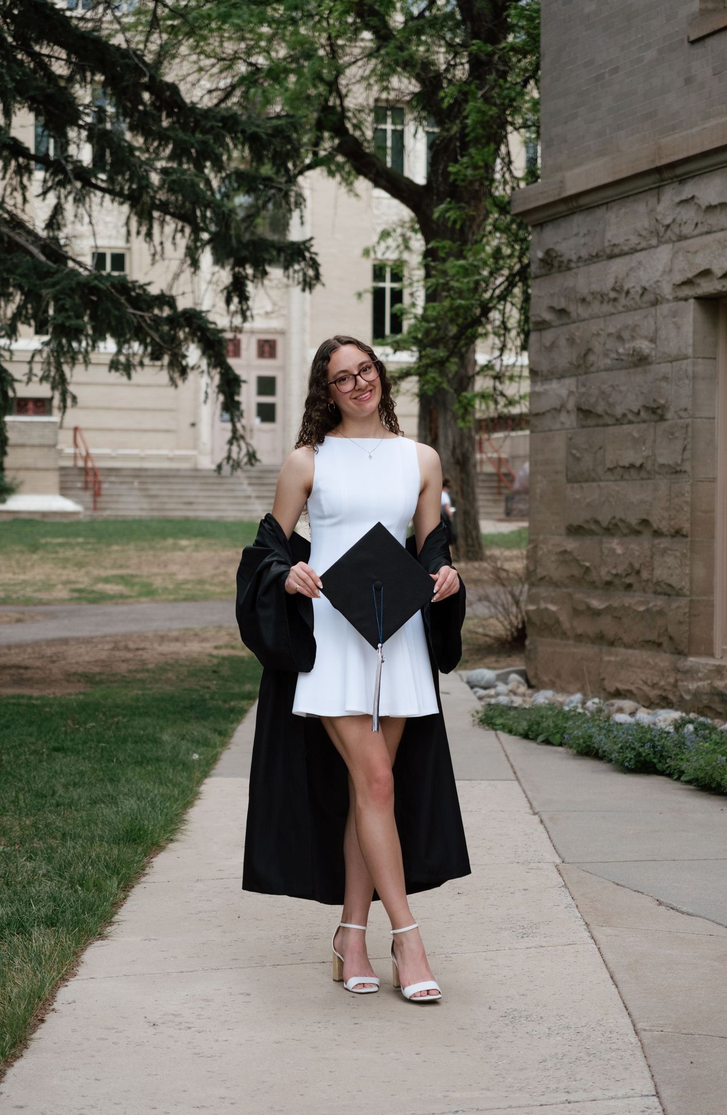 A young woman in a white dress holds graduation cap and gown outdoors on a sunny campus walkway.