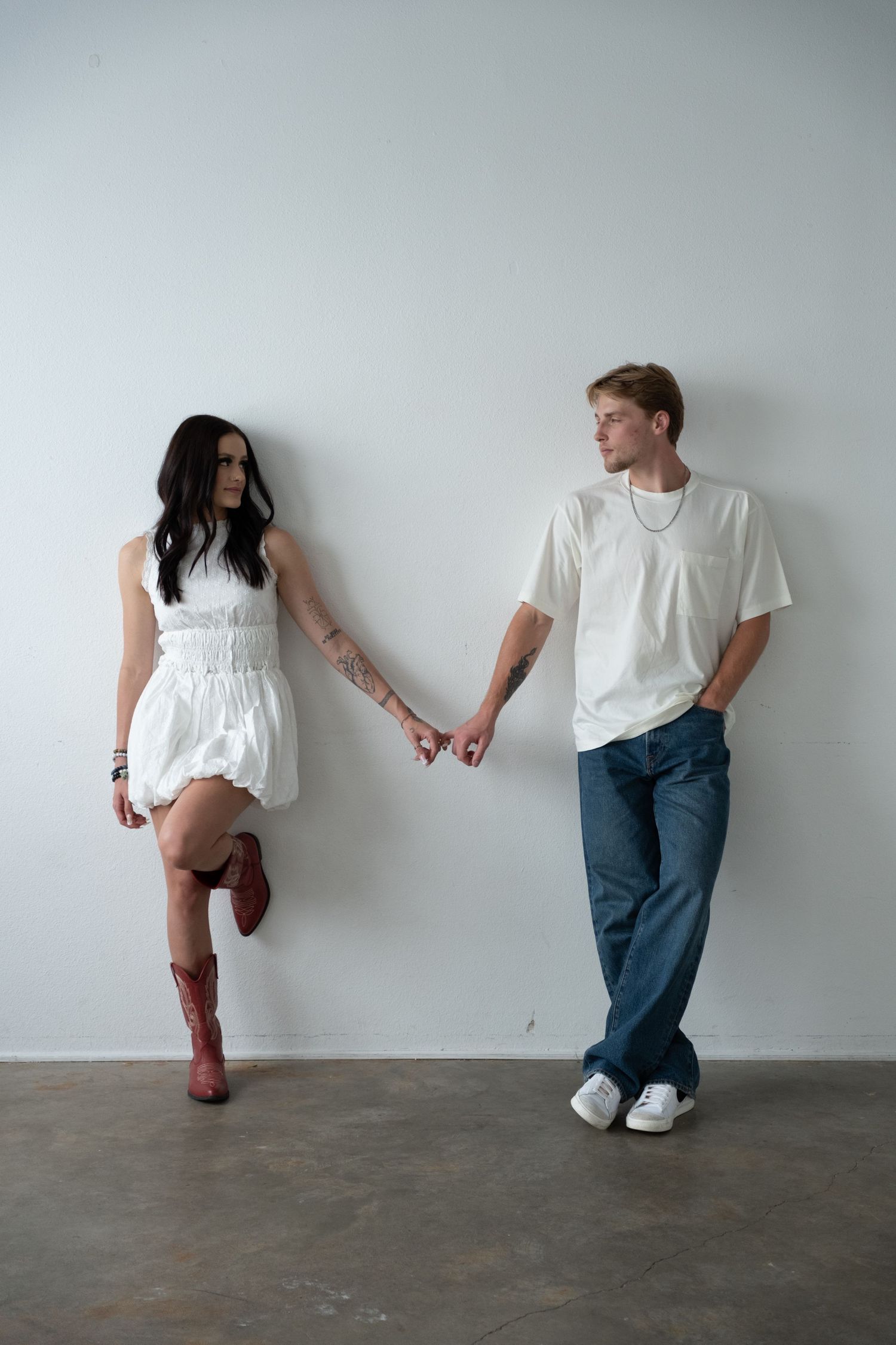 A young couple in white outfits holds hands against a white wall, woman in mini dress and red boots.
