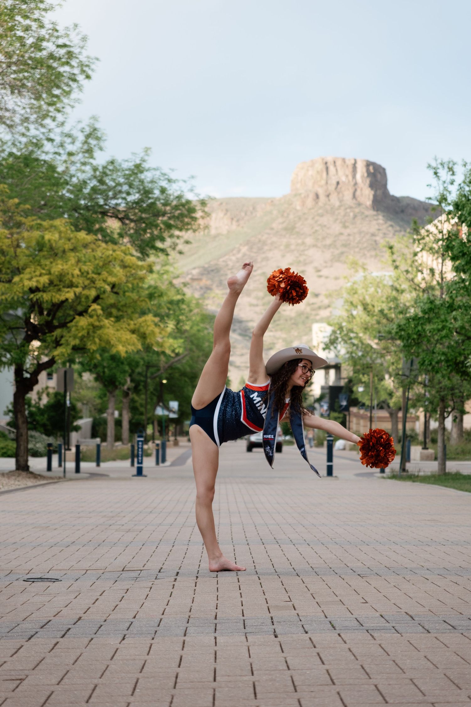 Cheerleader in cowboy hat performs a high kick with pom-poms on a brick path with mountain backdrop.