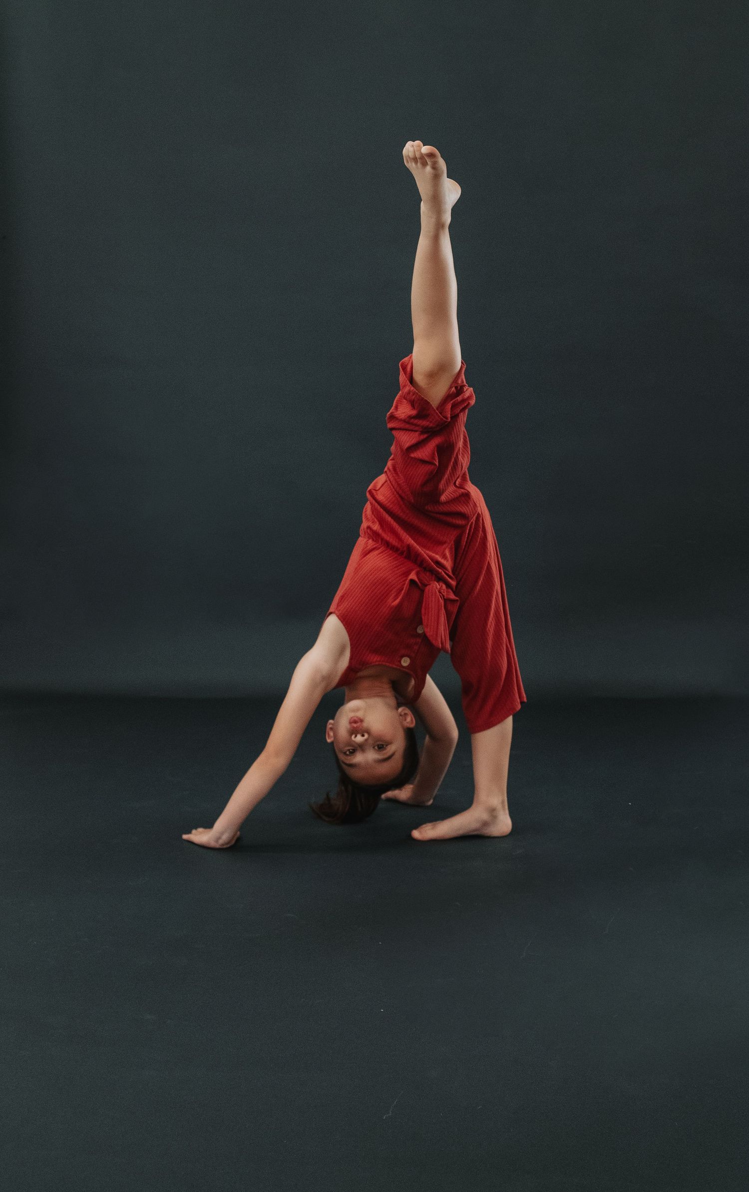 Young dancer in red outfit performs one-handed cartwheel against dark background.