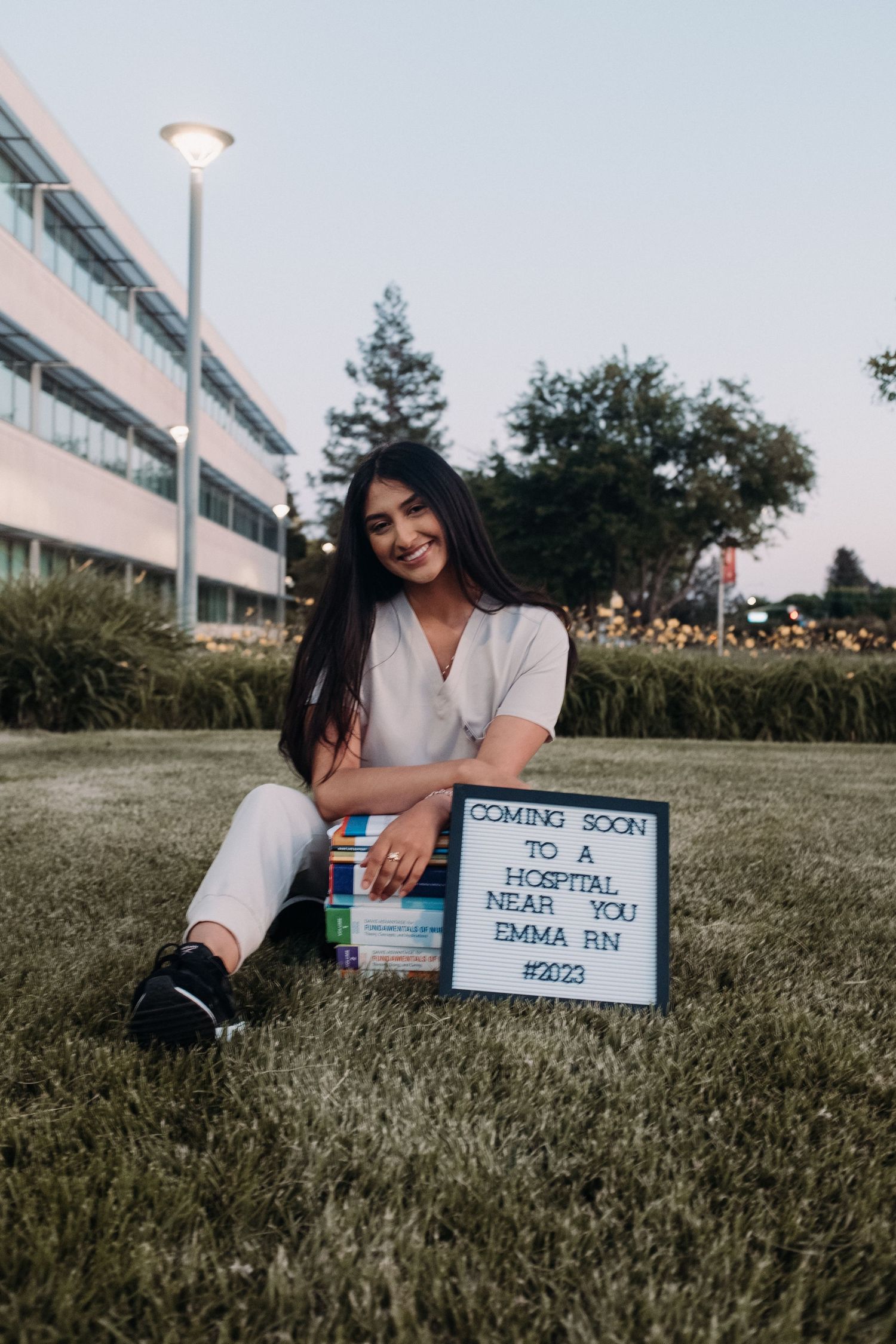 Graduate in white outfit sitting on grass with celebratory sign.