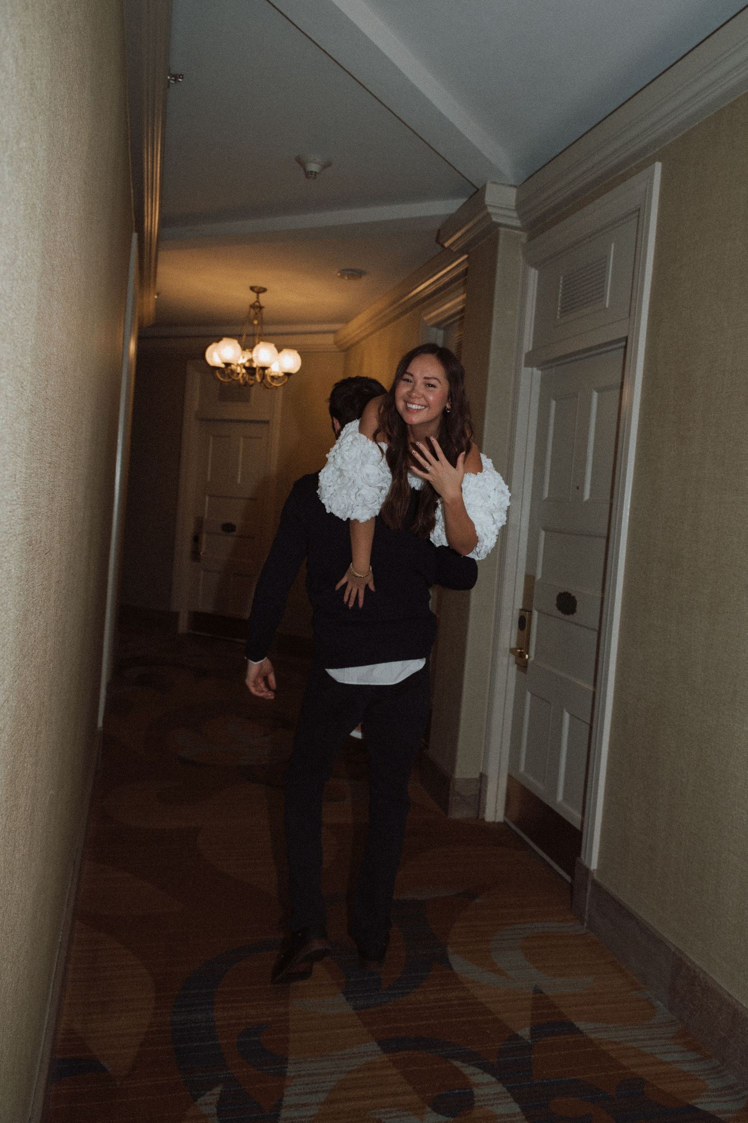 Smiling bride in white dress carried by groom through elegant hotel hallway with chandelier lighting.