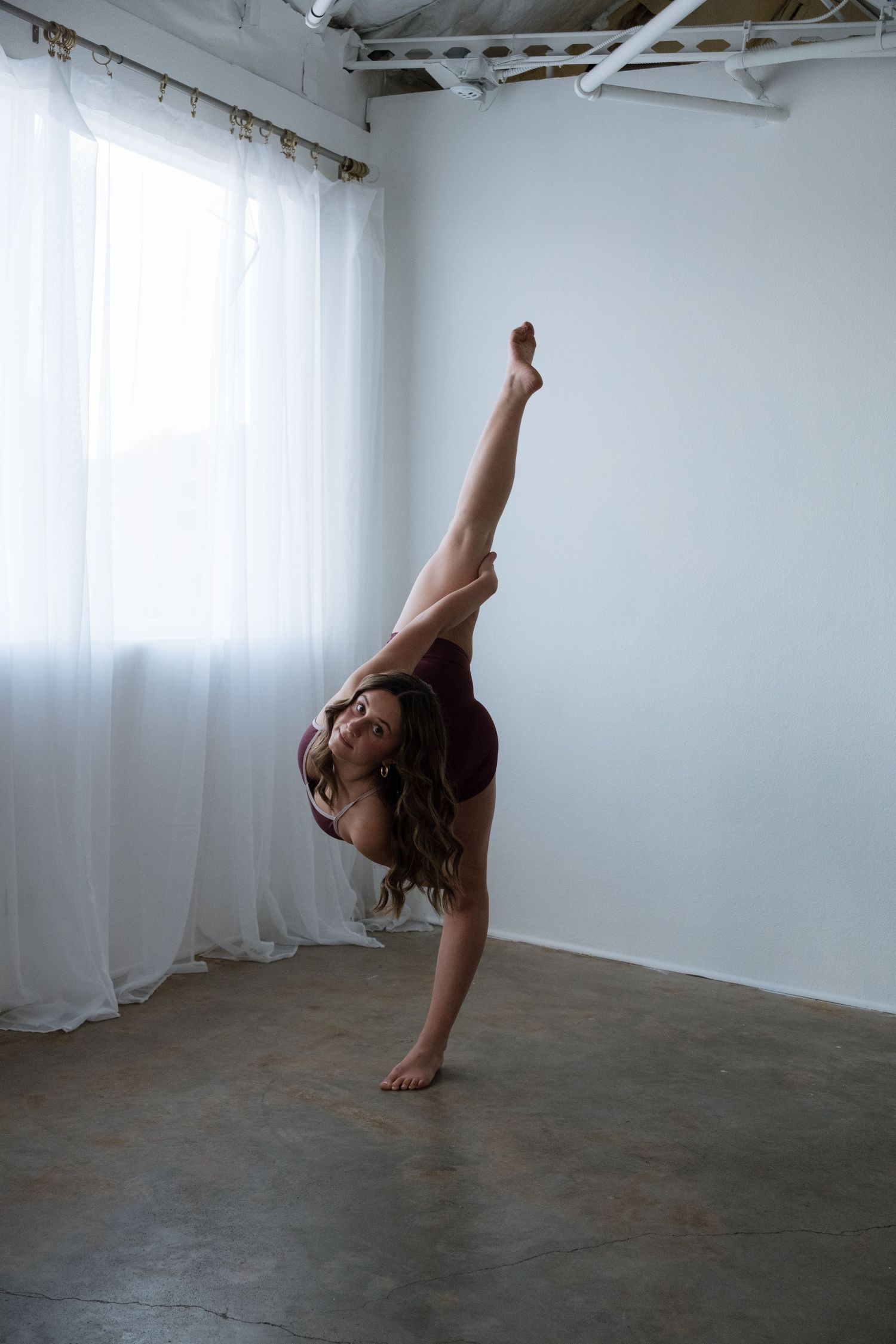 Flexible dancer in burgundy leotard performing a standing split in a bright white studio with sheer curtains.