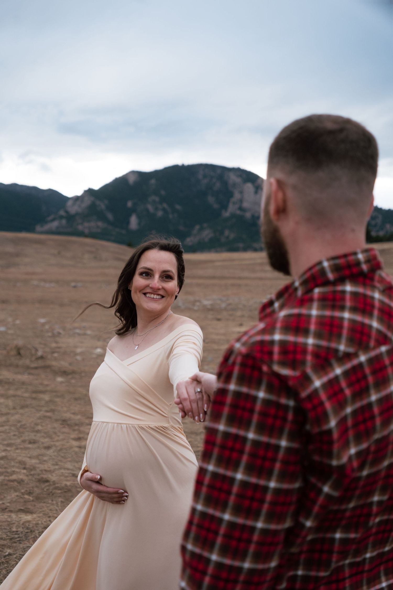 Pregnant woman in cream dress holds hands with bearded man in plaid shirt, smiling in front of mountain landscape.