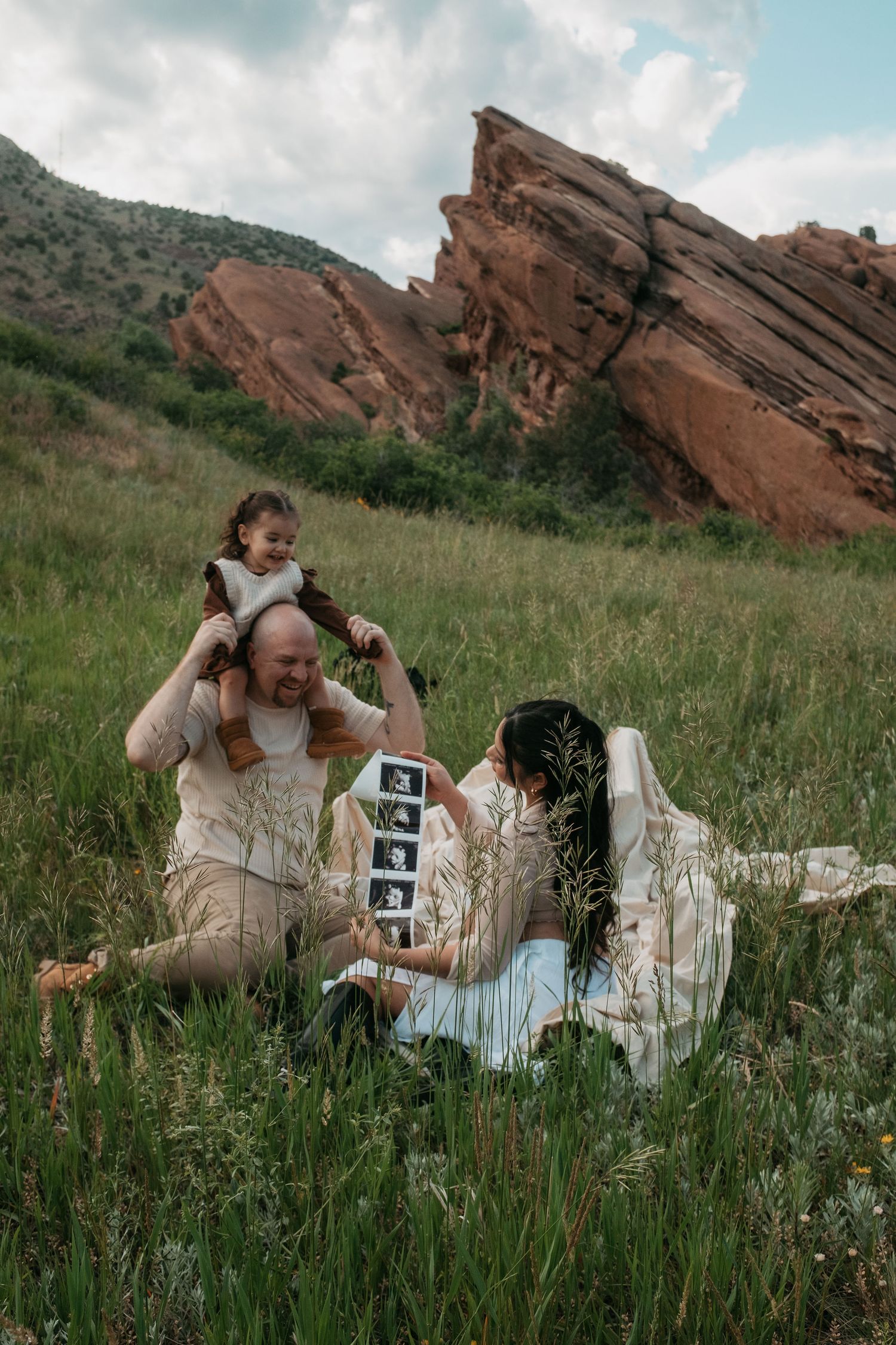 Family maternity announcement at Red Rocks Park, showing ultrasound photos in green meadow.